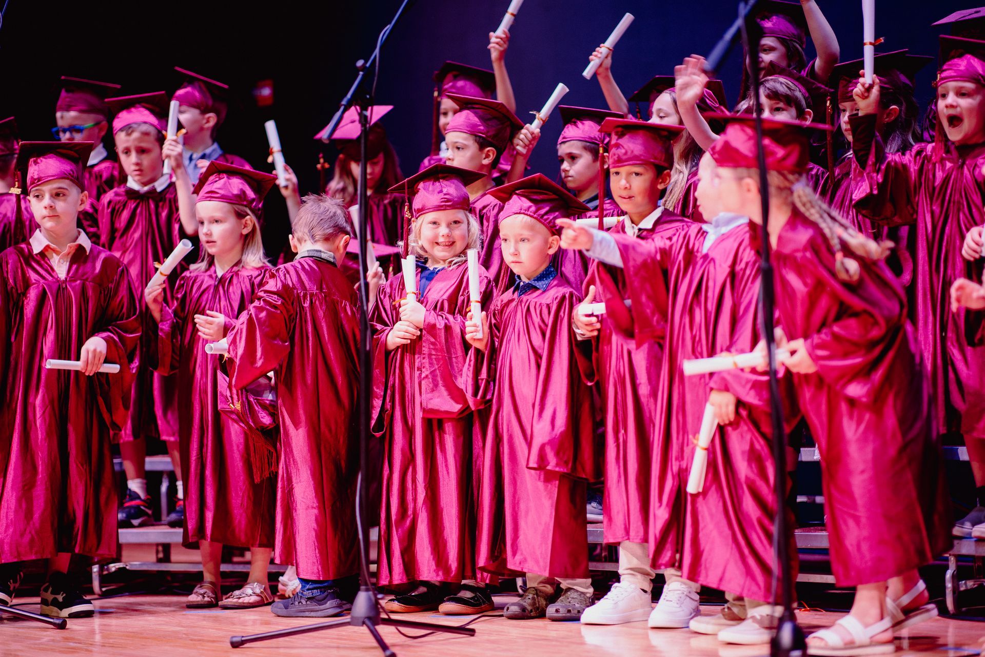 Children in pink graduation gowns and caps singing on a stage with microphones