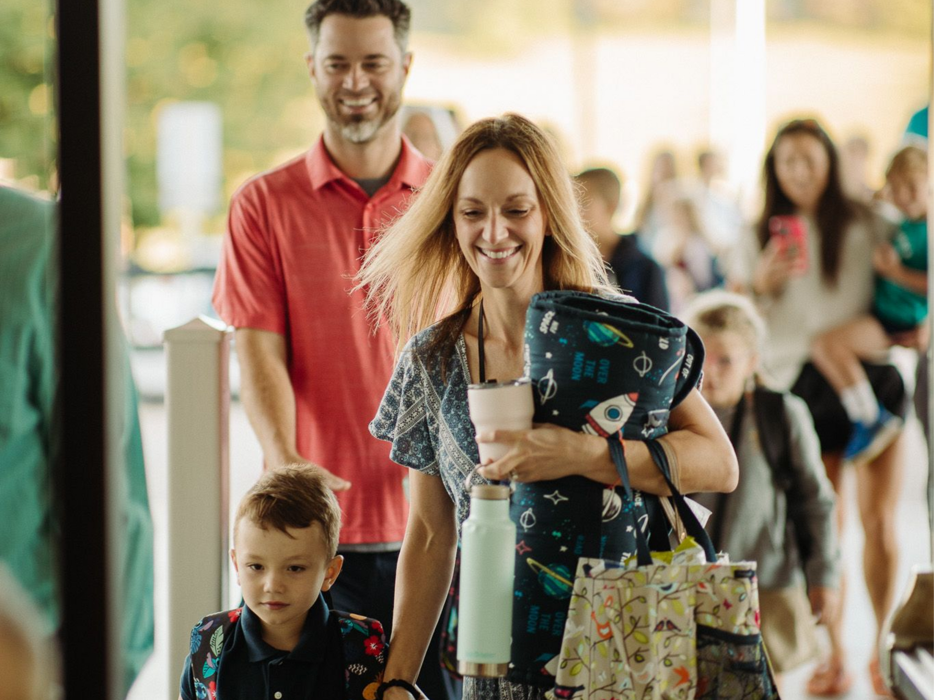 Family standing in a store aisle, smiling woman holding a drink and shopping bags, with a man and child behind her