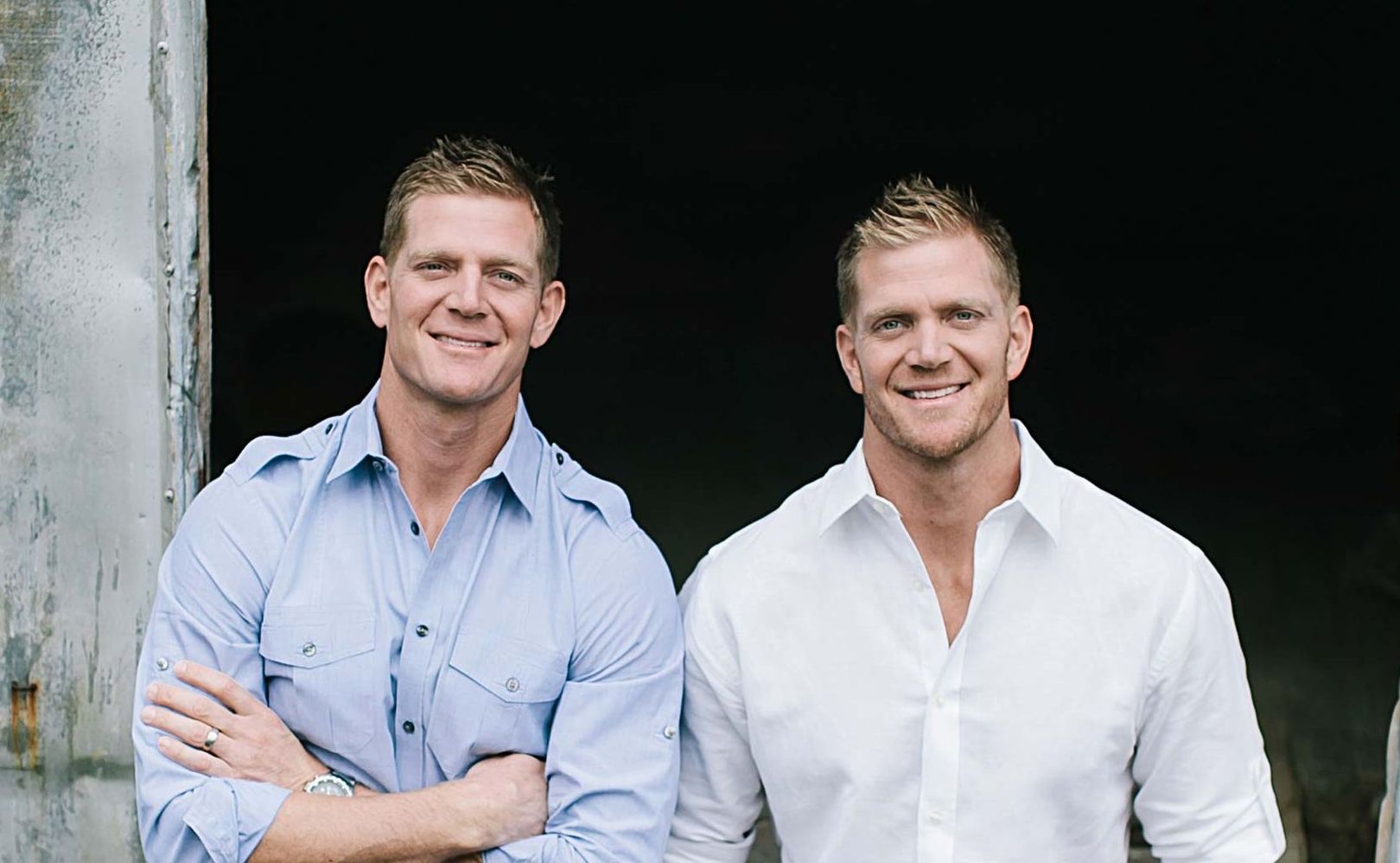 Two smiling men stand in front of a dark doorway, one in a light blue shirt and one in a white shirt.