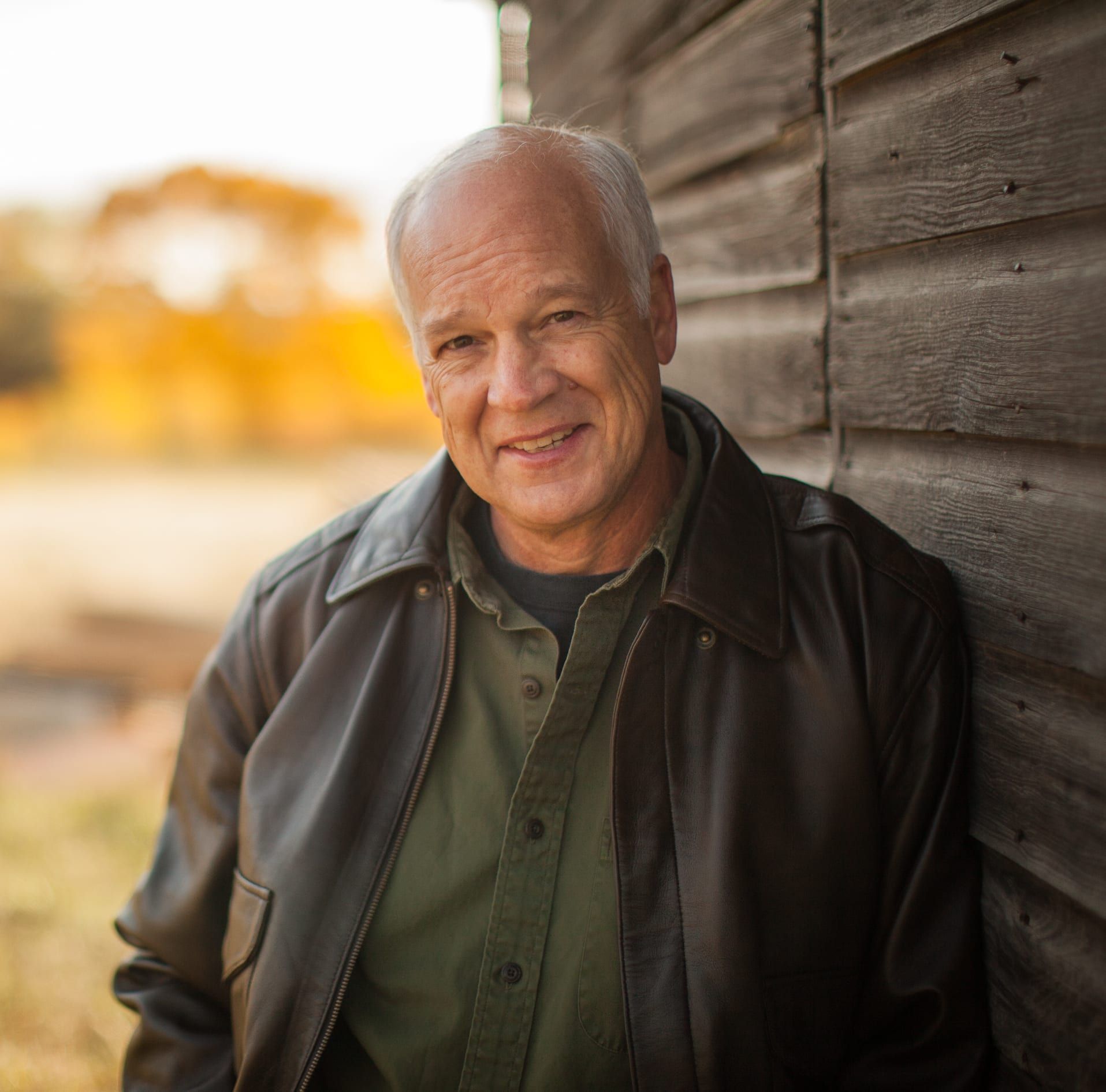 Man leaning against a wooden wall outdoors, wearing a dark leather jacket and smiling softly.