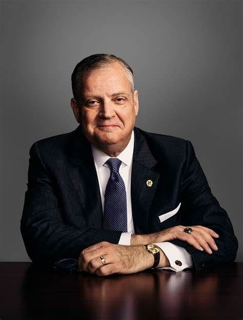 Portrait of a man in a dark suit seated at a table against a gray studio background.