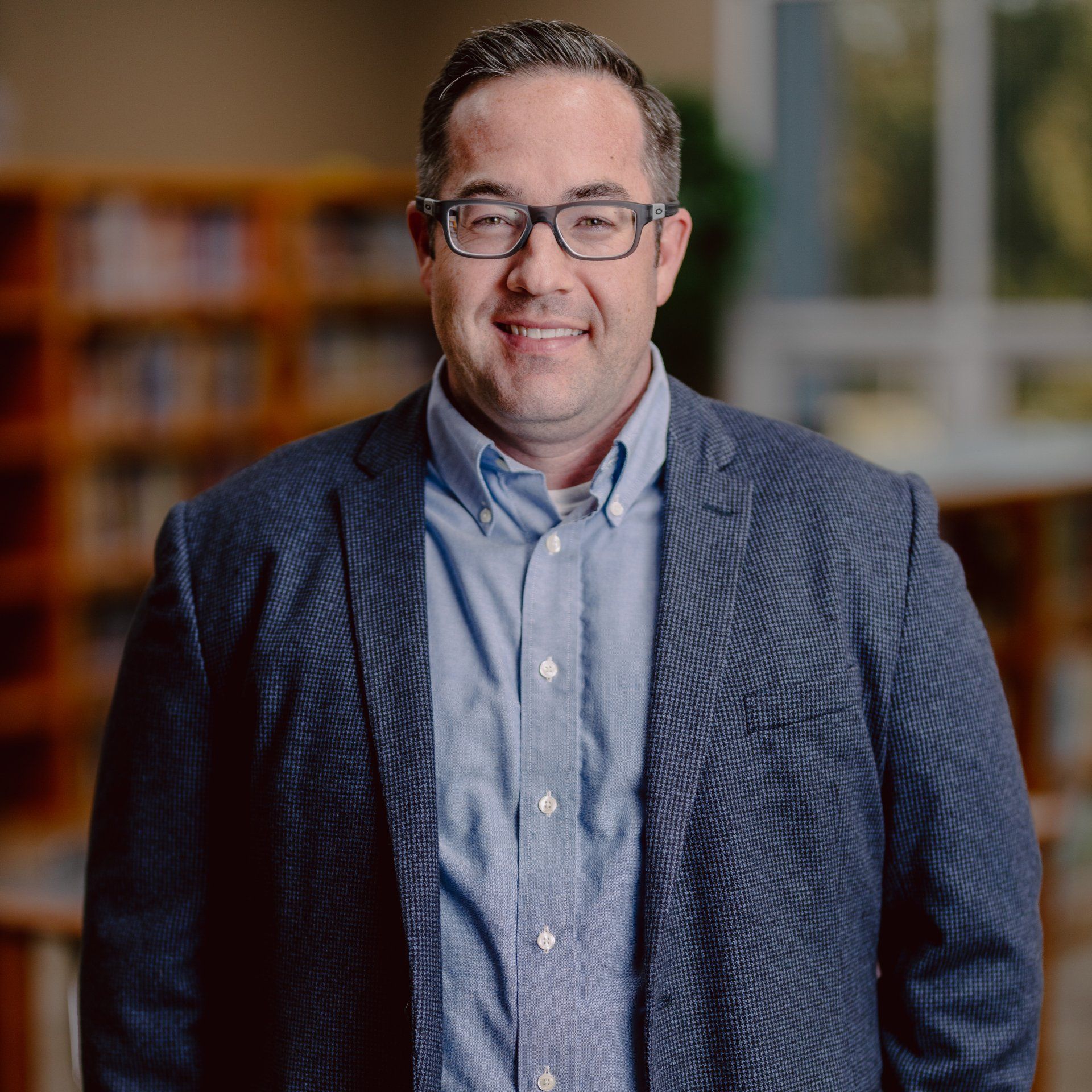Smiling man in glasses wearing a blue shirt and patterned blazer, standing indoors with a blurred bookshelf background