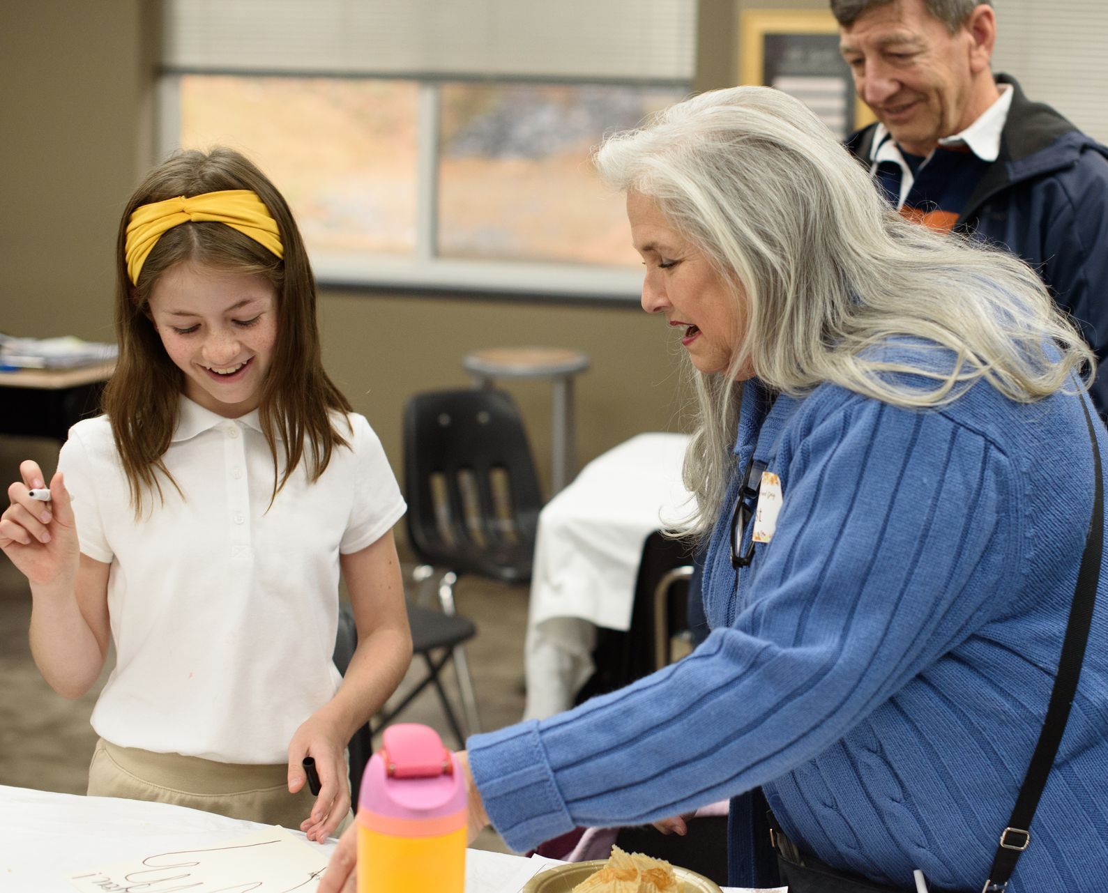 Two people smiling and working at a table with craft supplies in a classroom