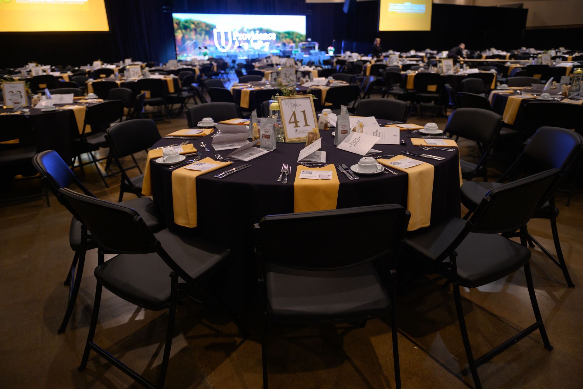 Round banquet tables set for an event in a dimly lit conference hall with a stage in the background