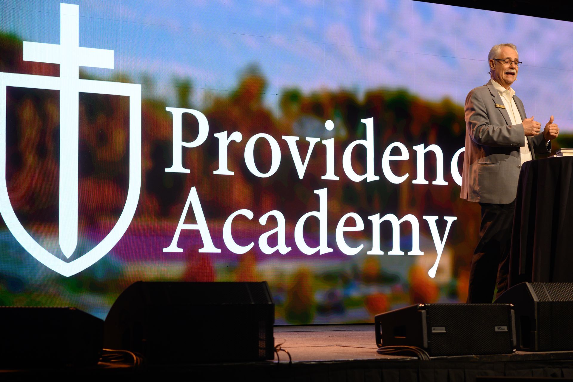Providence Academy logo on stage screen with a speaker at podium and cross emblem