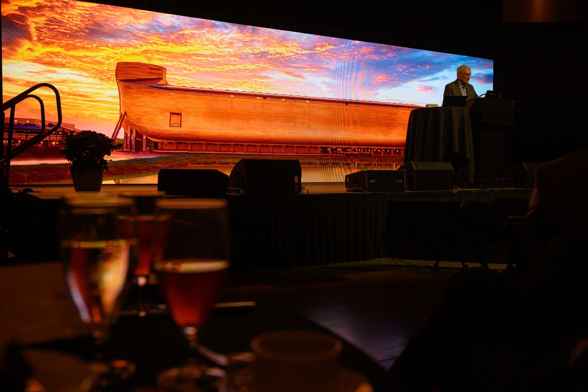 Large LED stage display of a boat at sunset, with silhouetted seating in the foreground