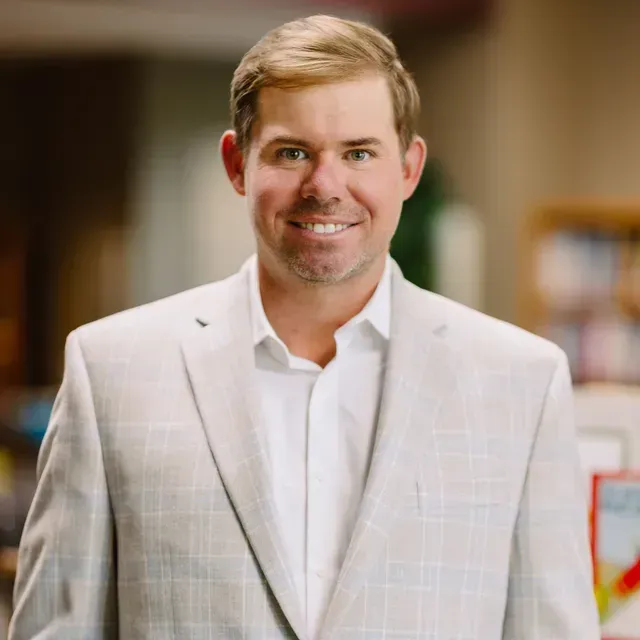 Smiling person in a light plaid blazer and white shirt, standing indoors with a blurred background.