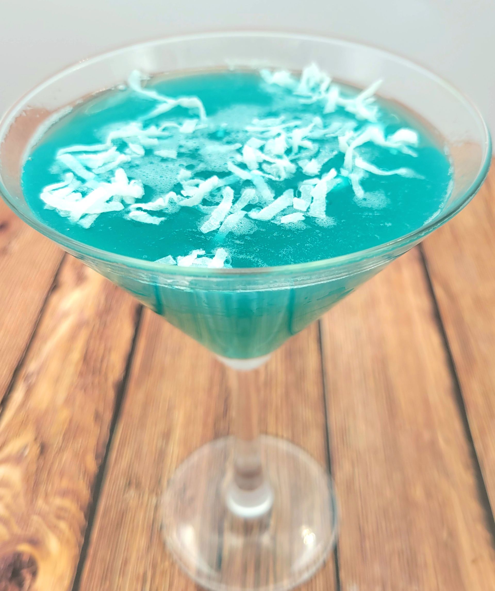 A martini glass filled with blue liquid and coconut on a wooden table.