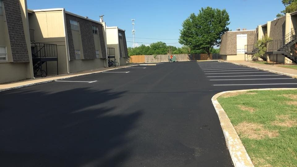 Empty apartment complex parking lot with fresh black asphalt, tan buildings, and a grassy curb under a clear sky