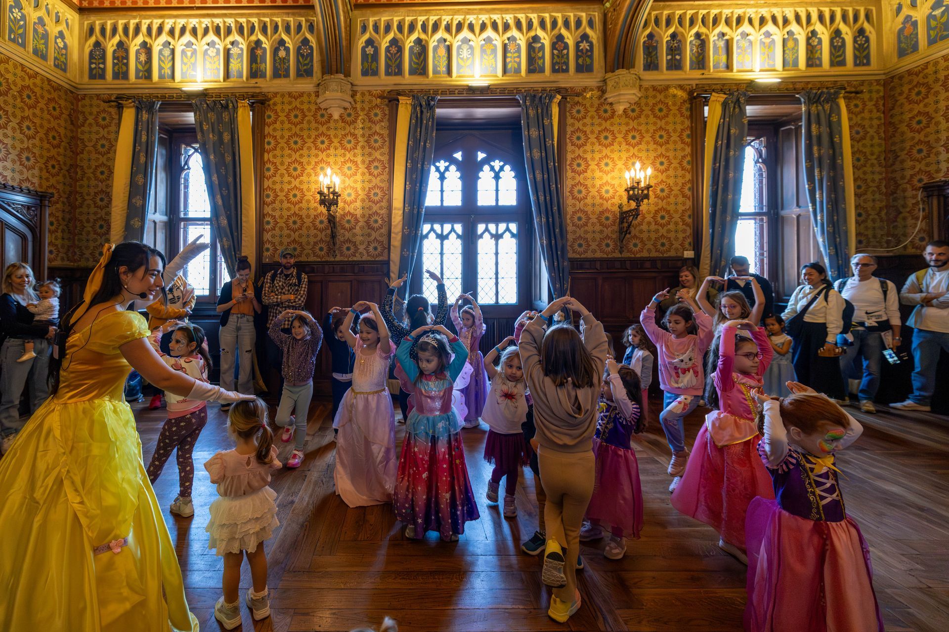 Una donna in abito da ballo giallo danza con un gruppo di bambini in costume all'interno di una sala riccamente decorata e rivestita di pannelli di legno.