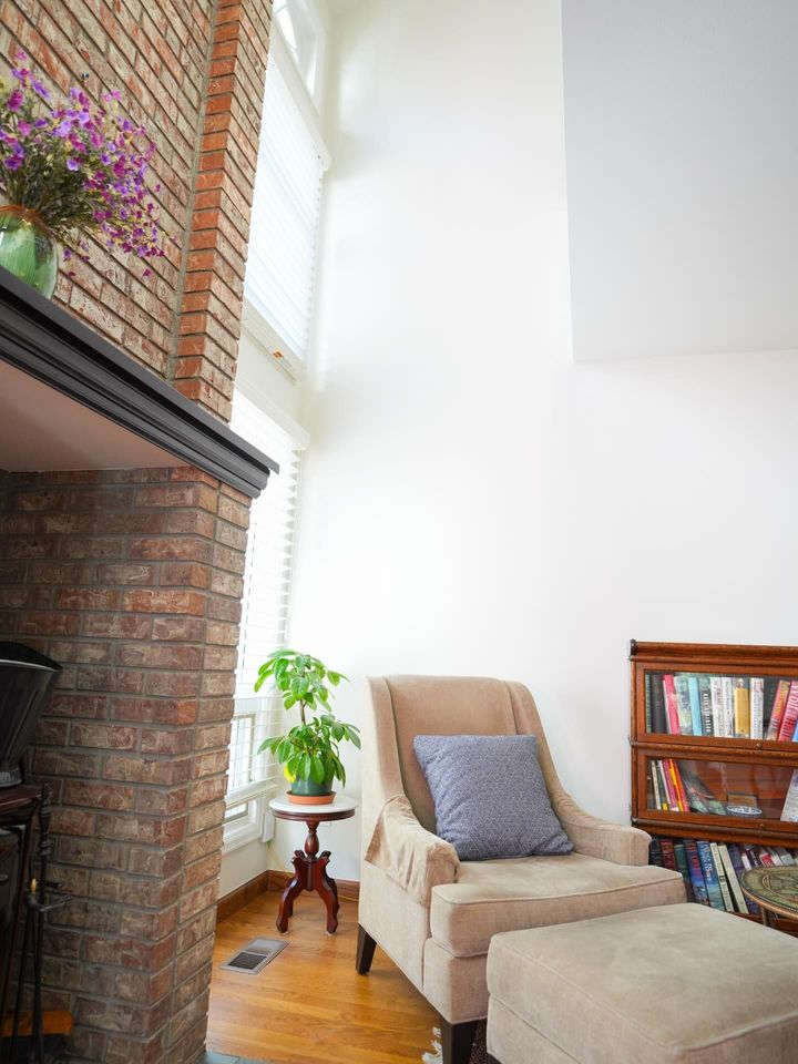 Bright living room corner with beige armchair, ottoman, bookshelf, brick fireplace, and potted plant
