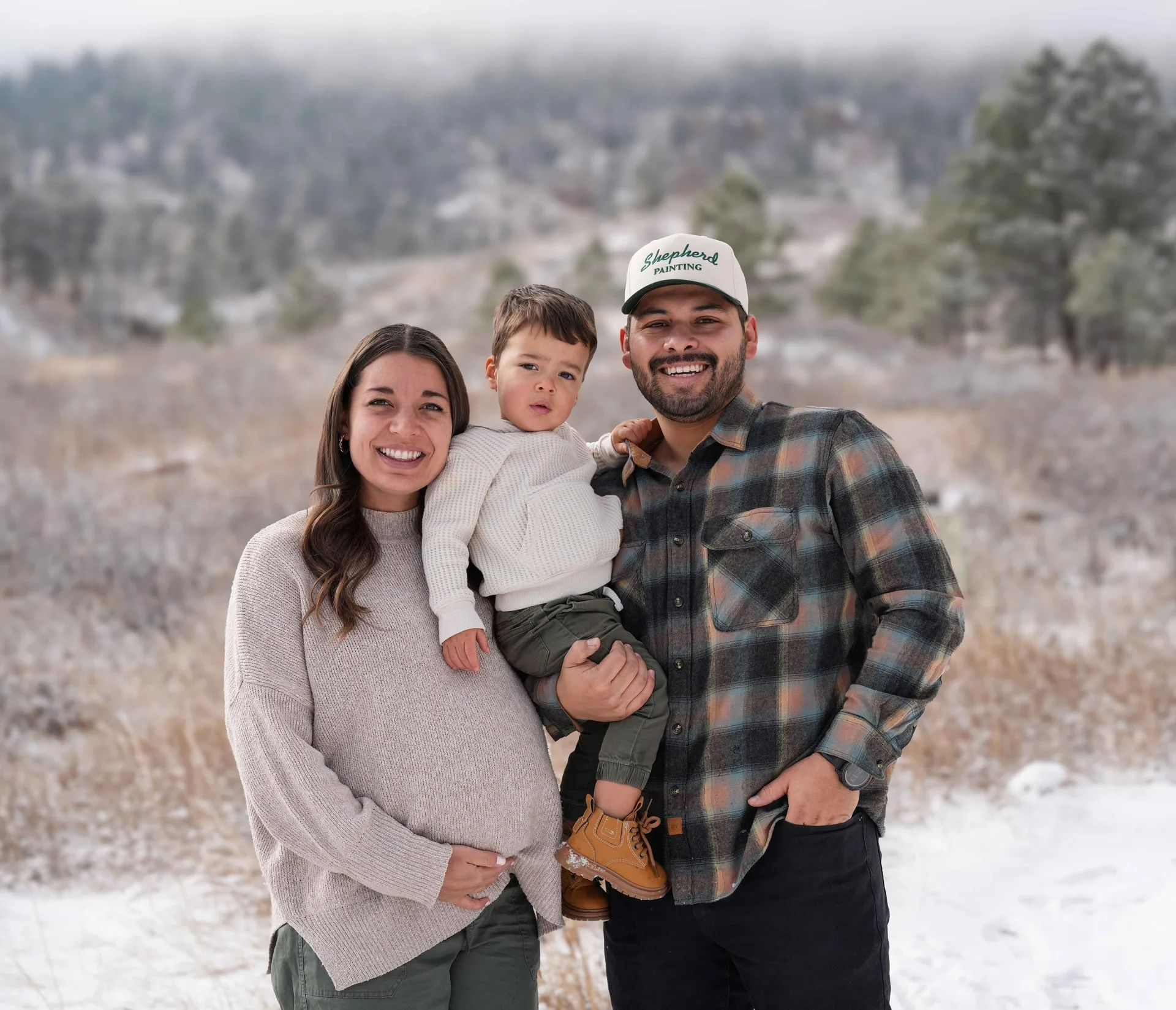 A family of three smiling outdoors in a snowy field, with trees and mountains in the background.