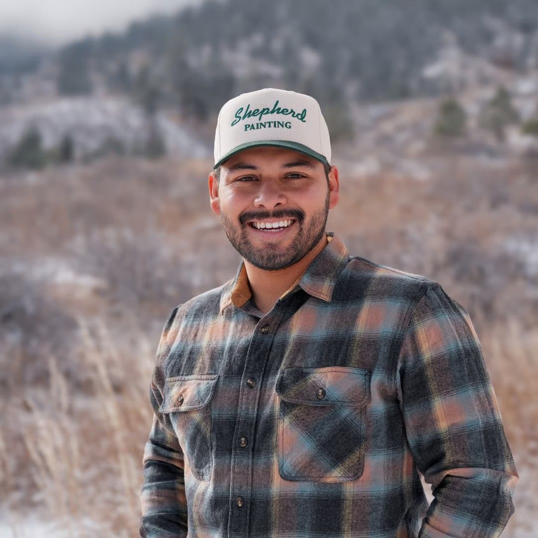 Smiling person in a plaid shirt and cap standing outdoors in a snowy mountain landscape