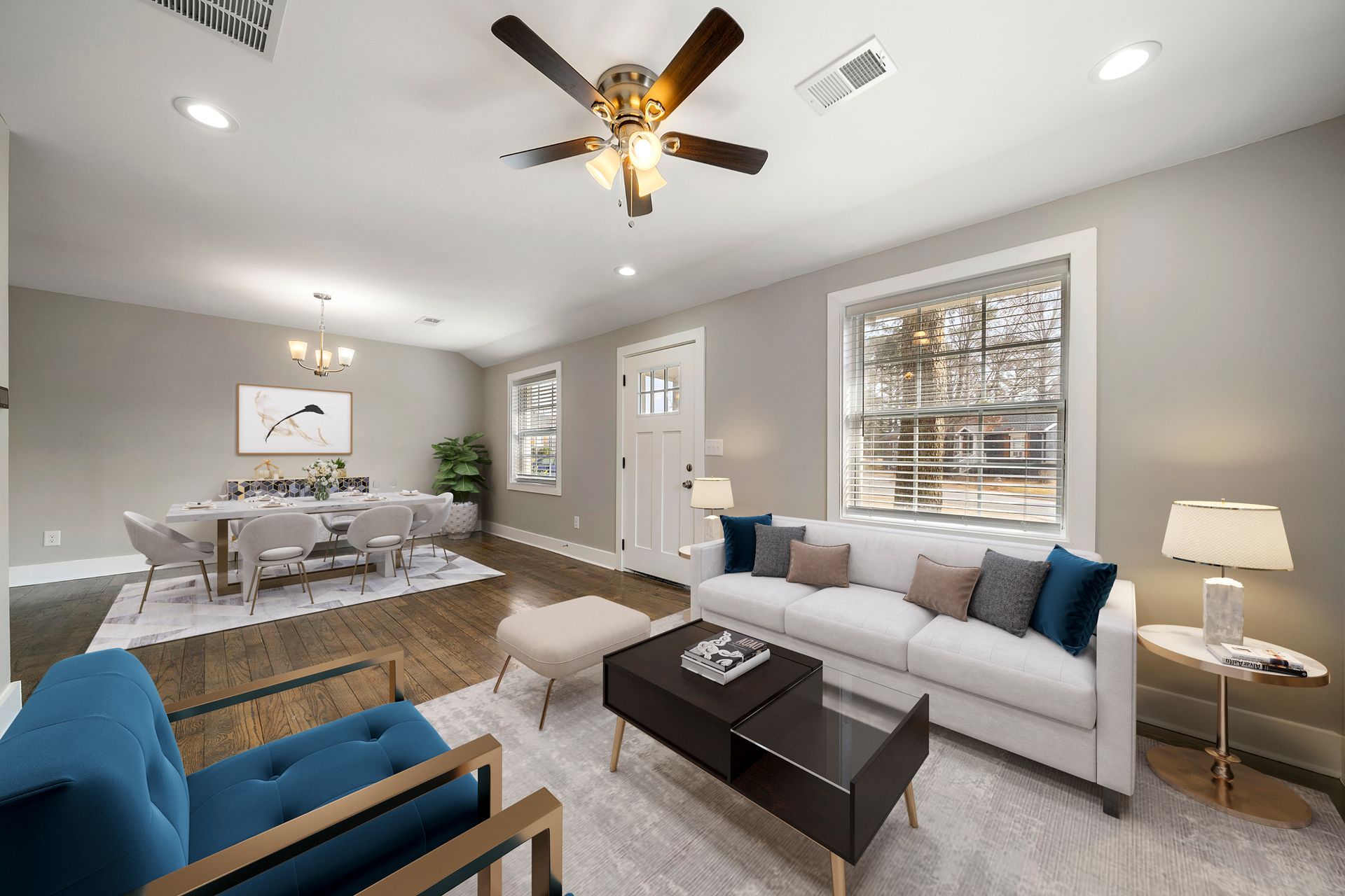 Bright living room with white sofa, blue chair, coffee table, dining area, ceiling fan, and large windows.