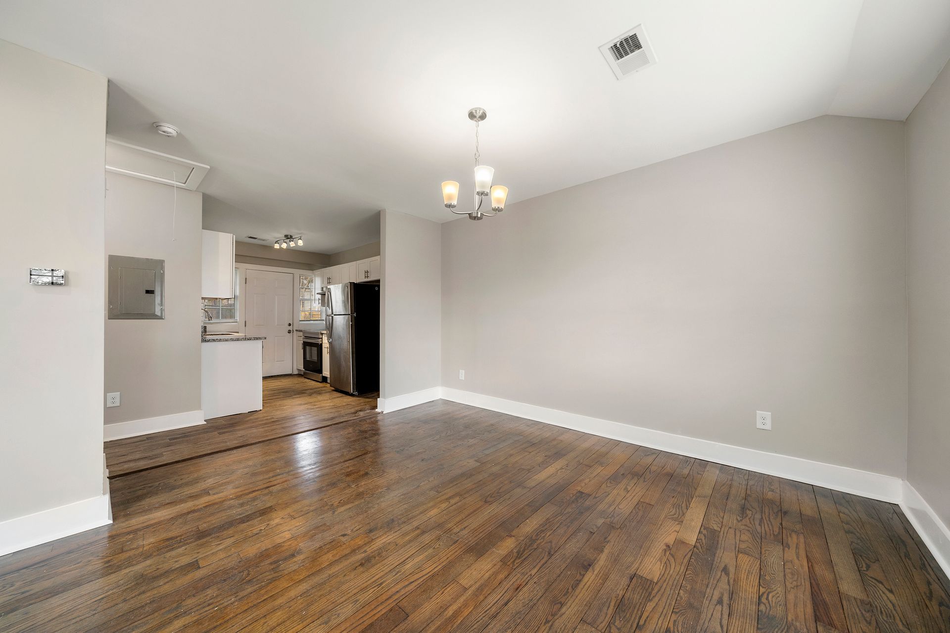 Empty living room with hardwood floors, gray walls, and a view into the kitchen.