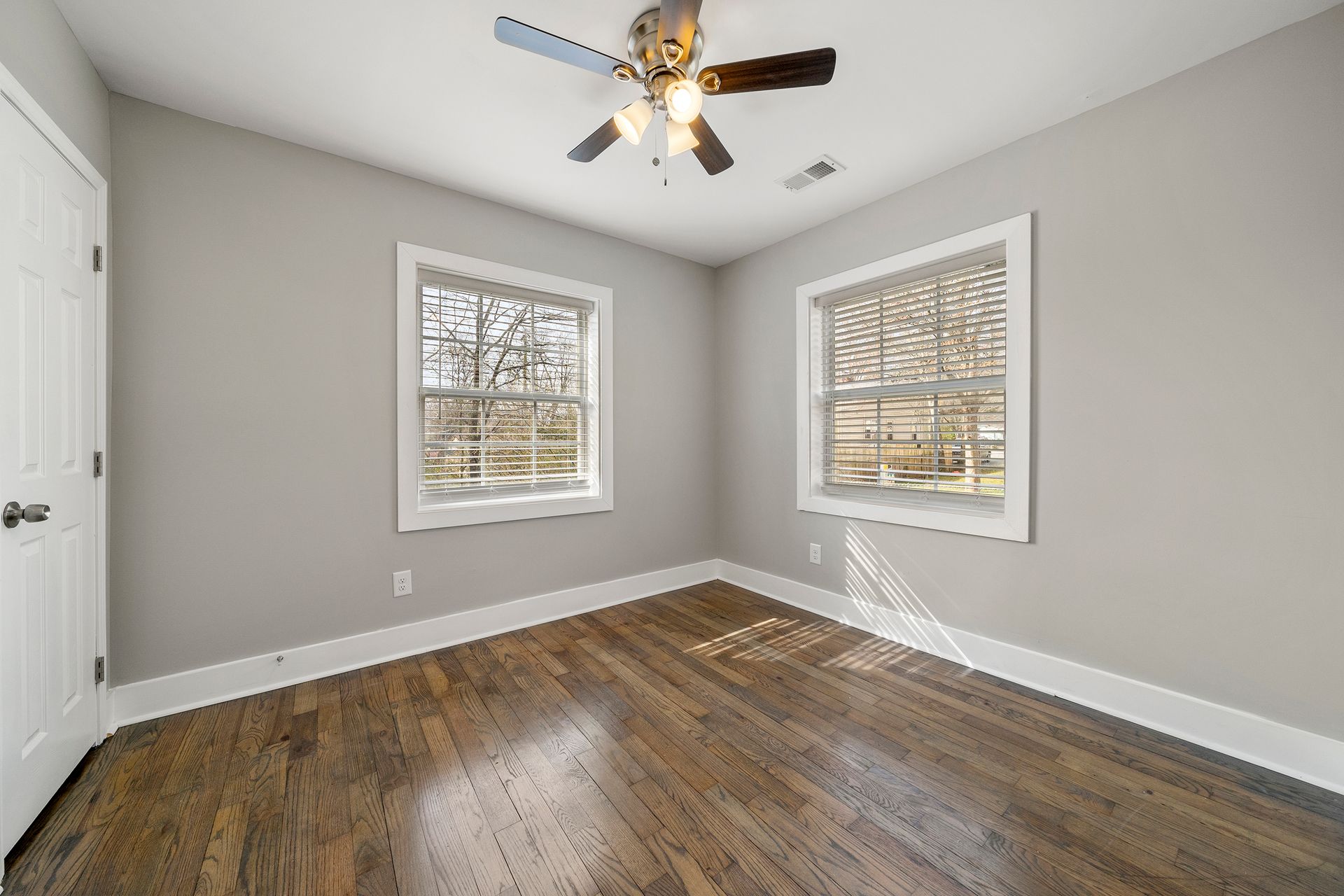 Empty gray bedroom with hardwood floors, two windows, and a ceiling fan with lights