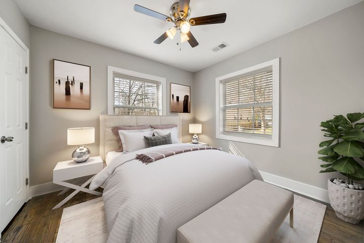 Cozy beige bedroom with a bed, nightstands, lamps, ceiling fan, windows, and framed wall art.