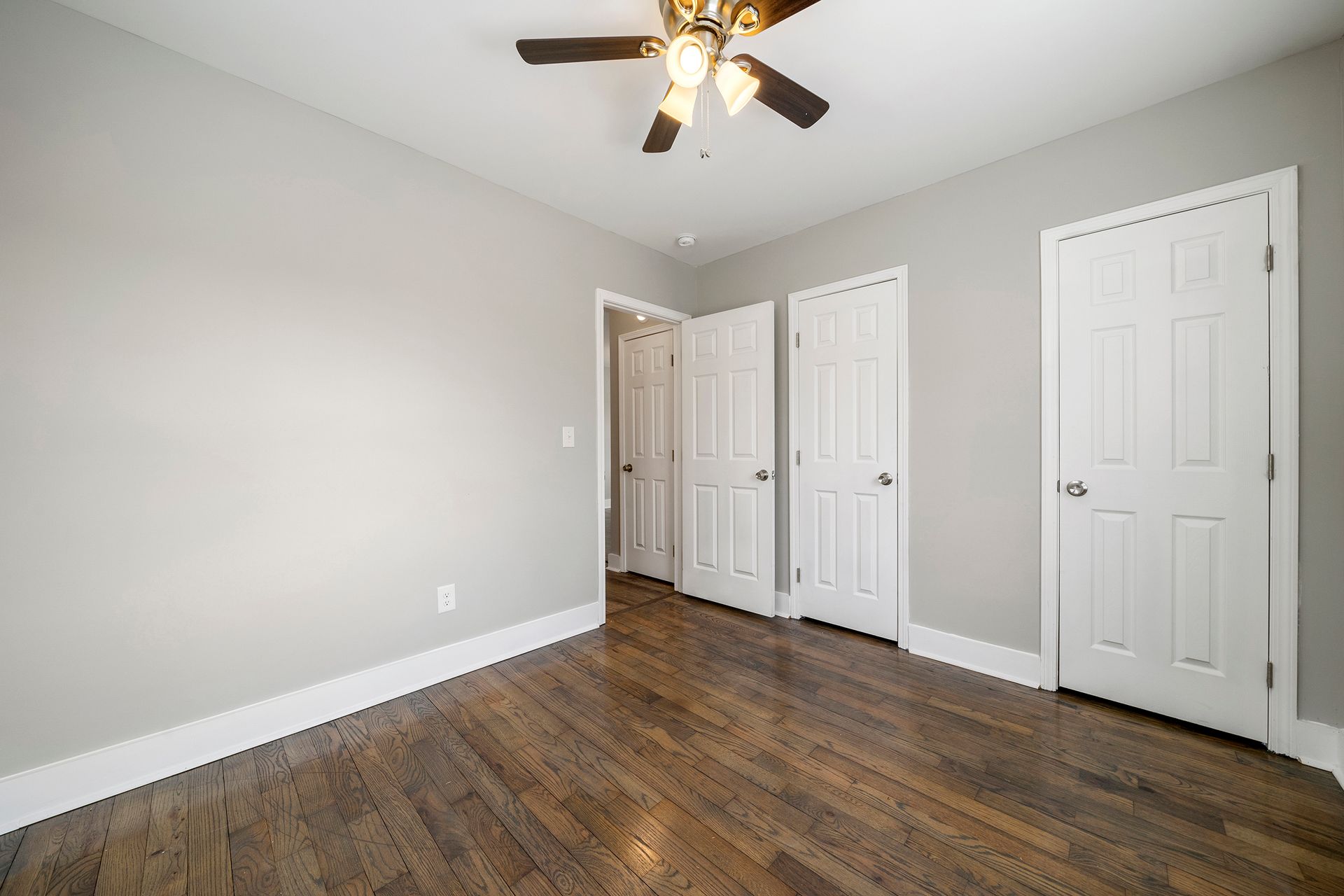Empty bedroom with gray walls, wood flooring, ceiling fan, and two white closet doors.