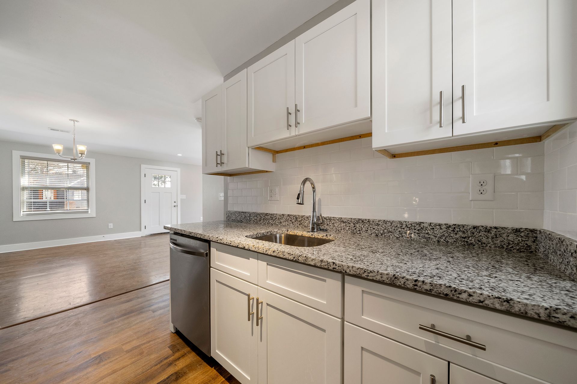 Bright kitchen with white cabinets, granite countertop, stainless dishwasher, and hardwood floors opening to a living area