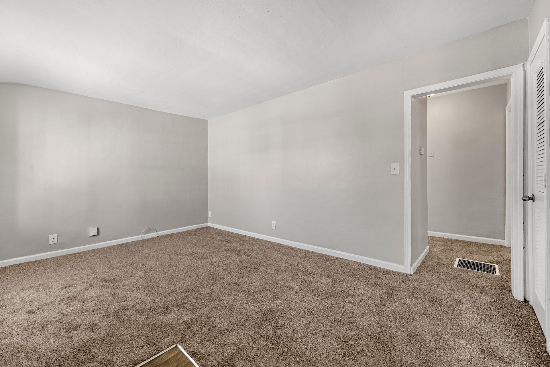 Empty beige-carpeted bedroom with light gray walls and an open doorway to a hall.