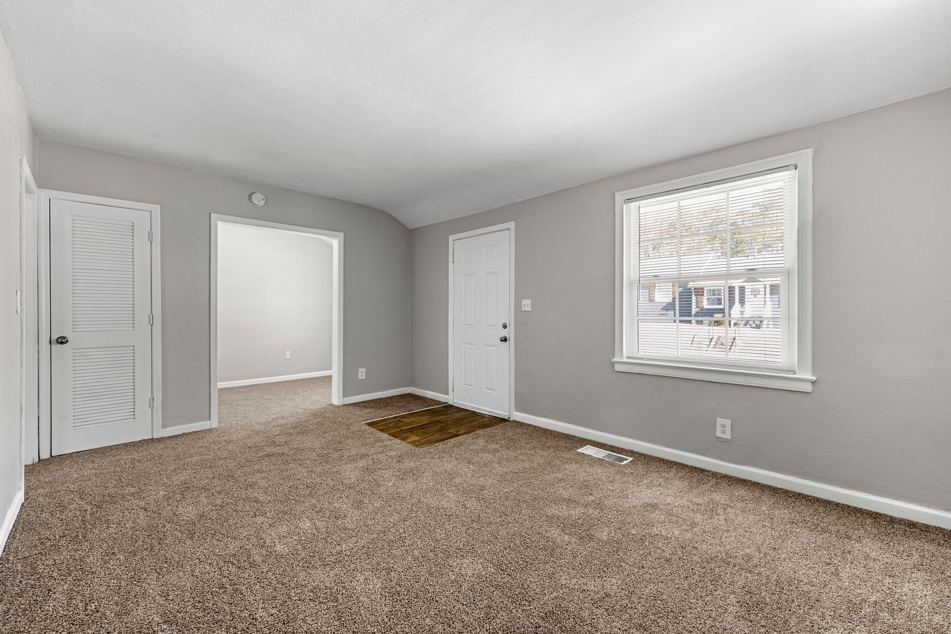 Empty beige-carpeted room with gray walls, white doors, and a window with blinds