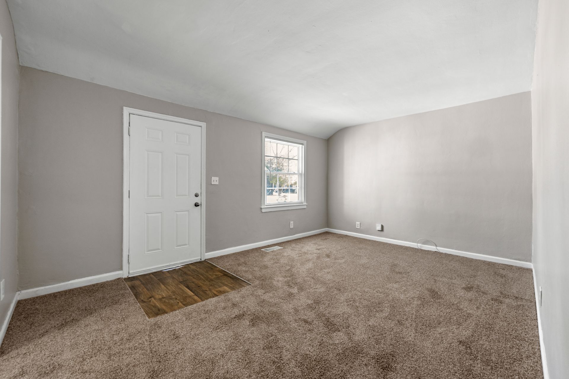 Empty beige bedroom with carpet, white door, and a small window on the left wall