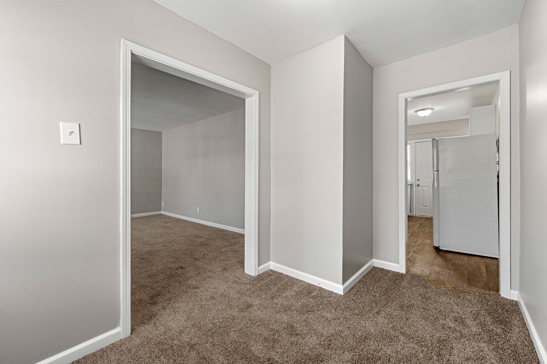 Empty gray hallway with carpet and white trim, showing two open doorways and a visible adjoining room.