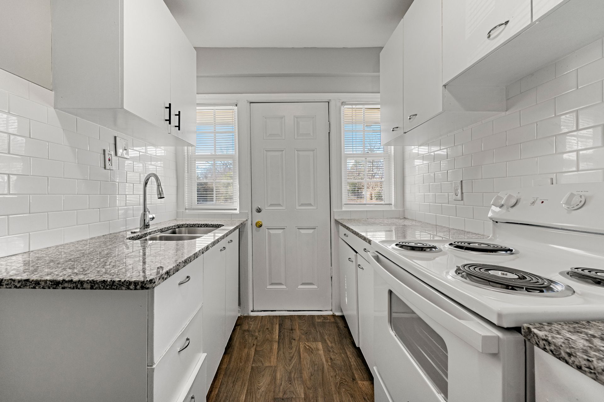 Bright narrow kitchen with white cabinets, subway tile, granite counters, and wood floor, viewed toward a back door