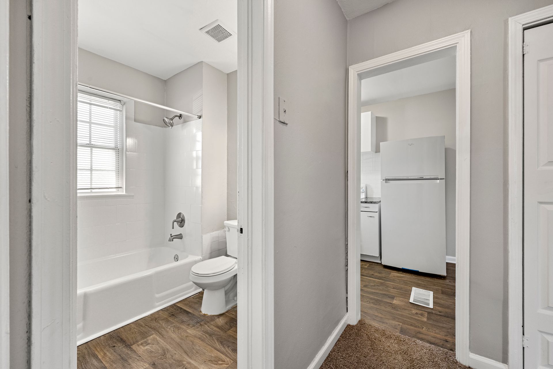 Small bathroom with white tub, toilet, and window beside a carpeted hallway and white refrigerator.