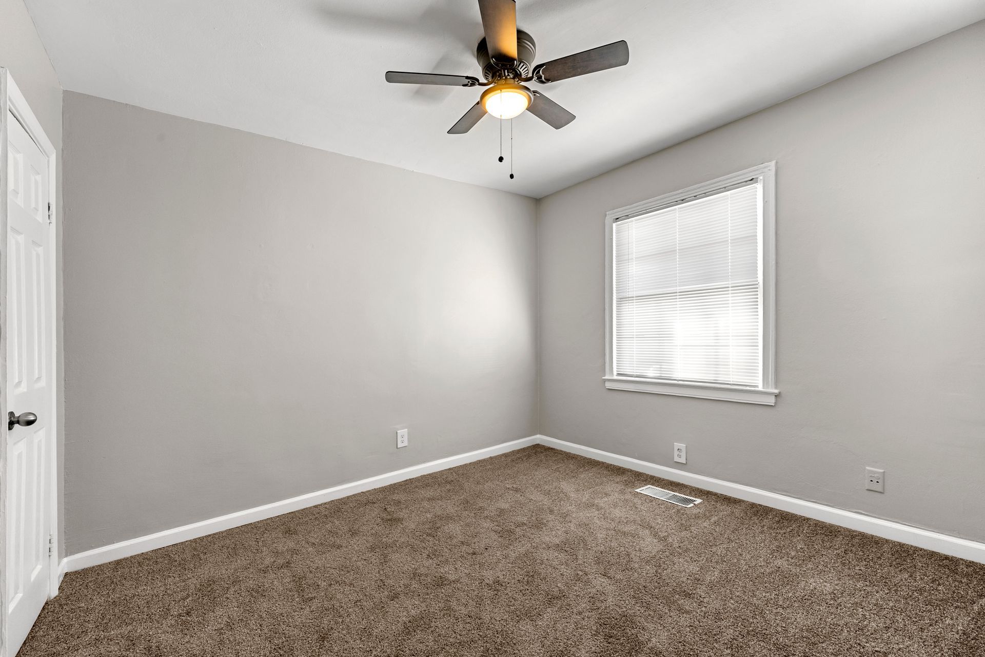 Empty gray carpeted bedroom with a ceiling fan and one window with blinds.