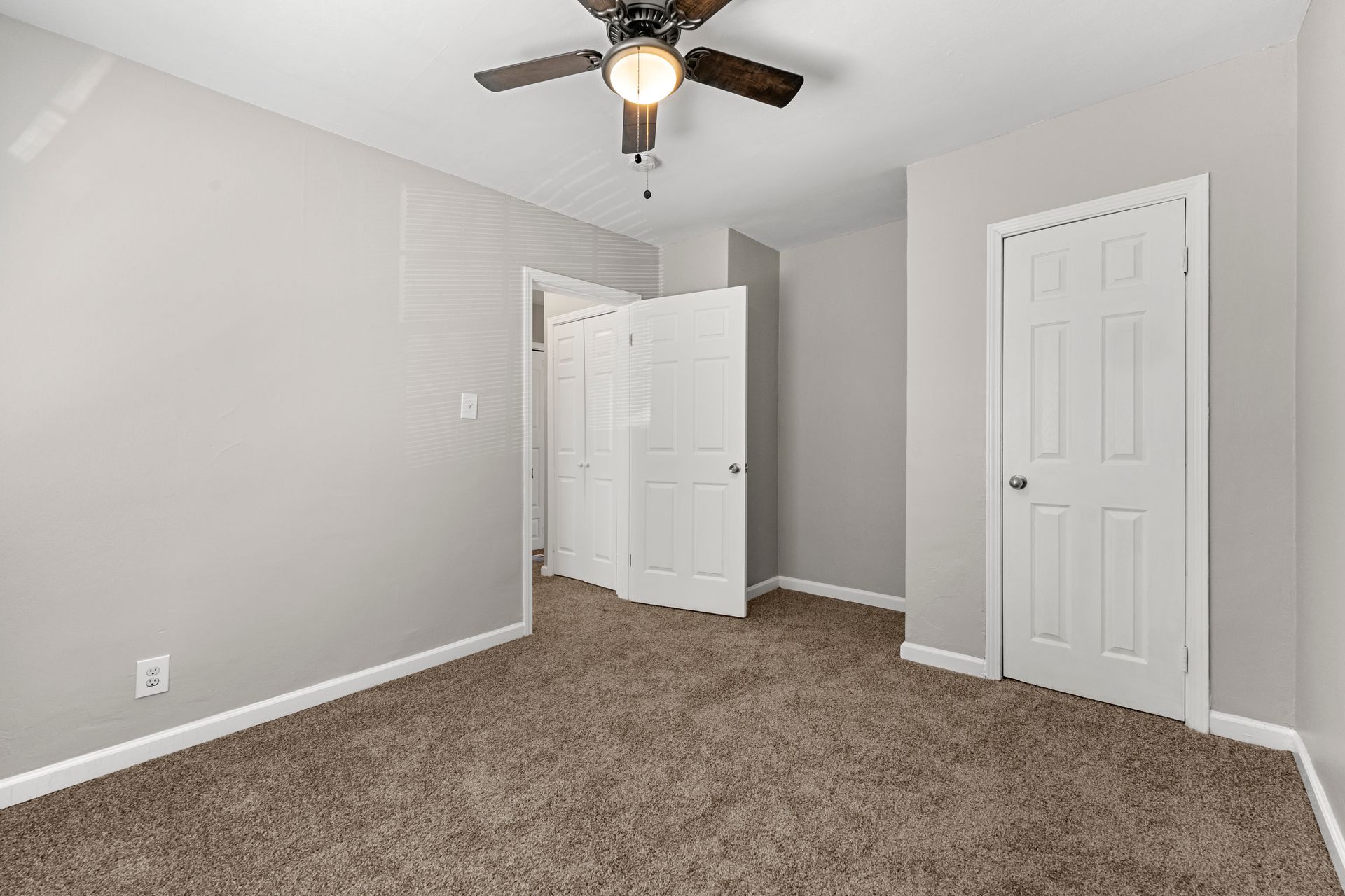 Empty beige bedroom with carpet, white closet doors, and a ceiling fan