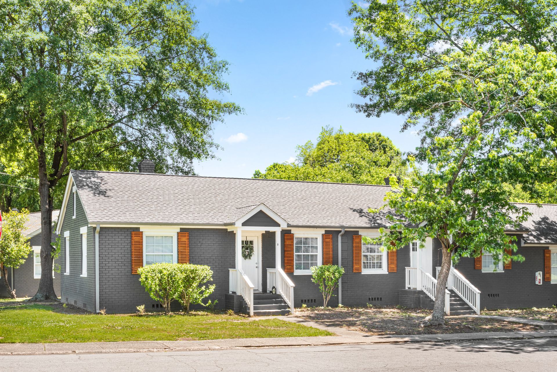 Single-story gray apartment building with white trim, orange shutters, and trees lining the street.