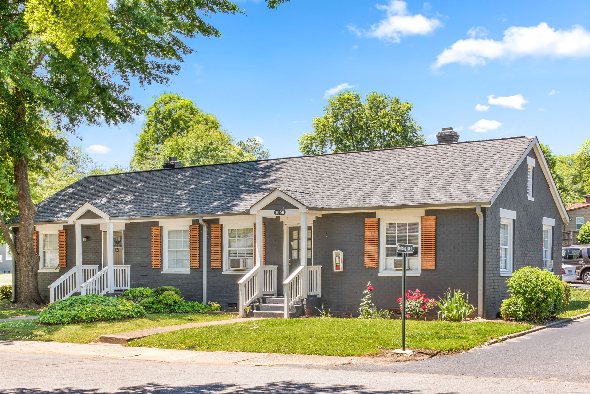 Gray house with white trim and orange shutters on a sunny street, surrounded by trees and greenery