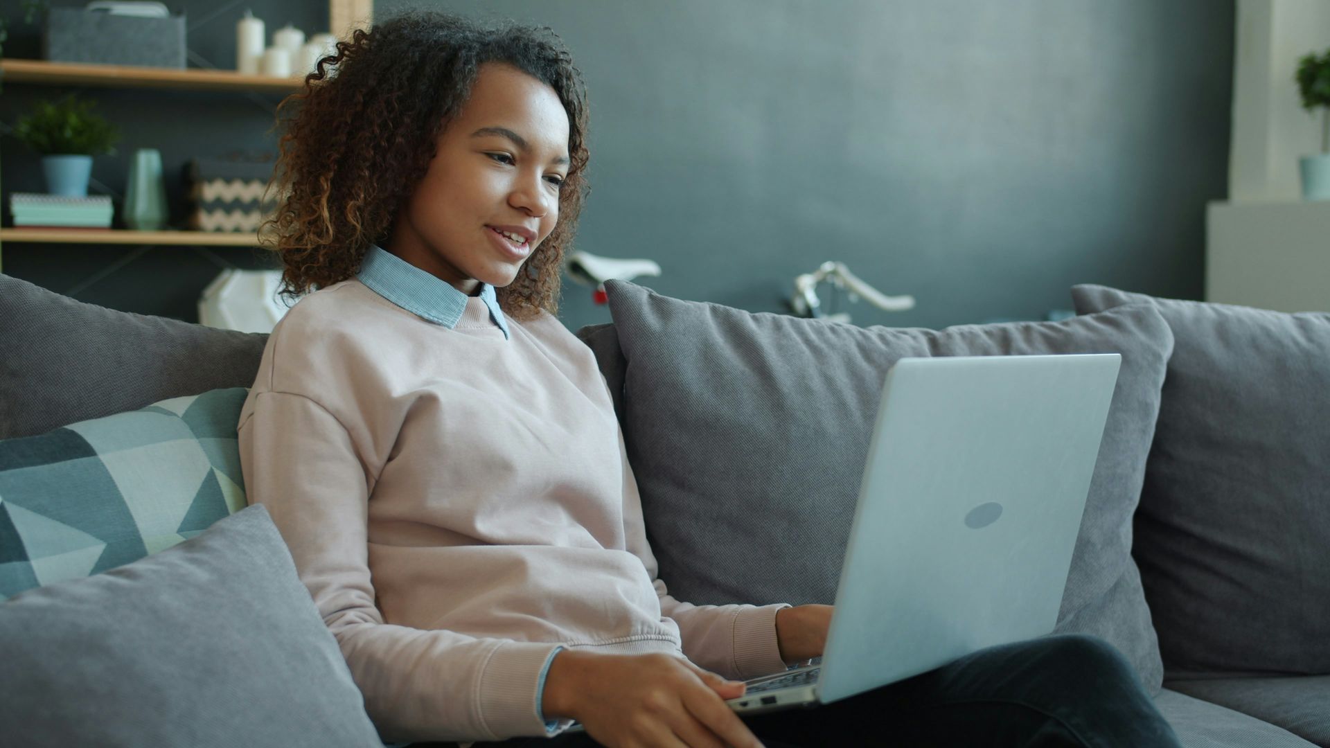 Woman using a laptop on a gray couch in a bright living room.
