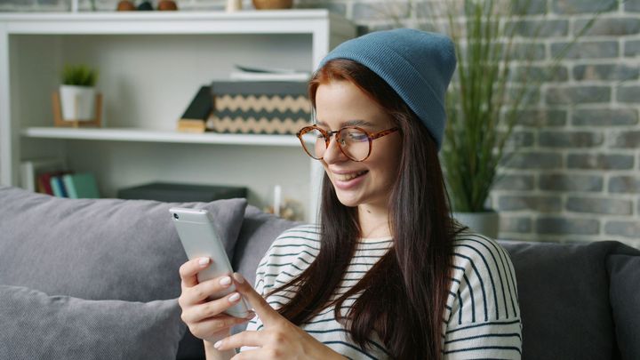 Person in glasses and a blue beanie smiling at a smartphone on a couch in a bright living room