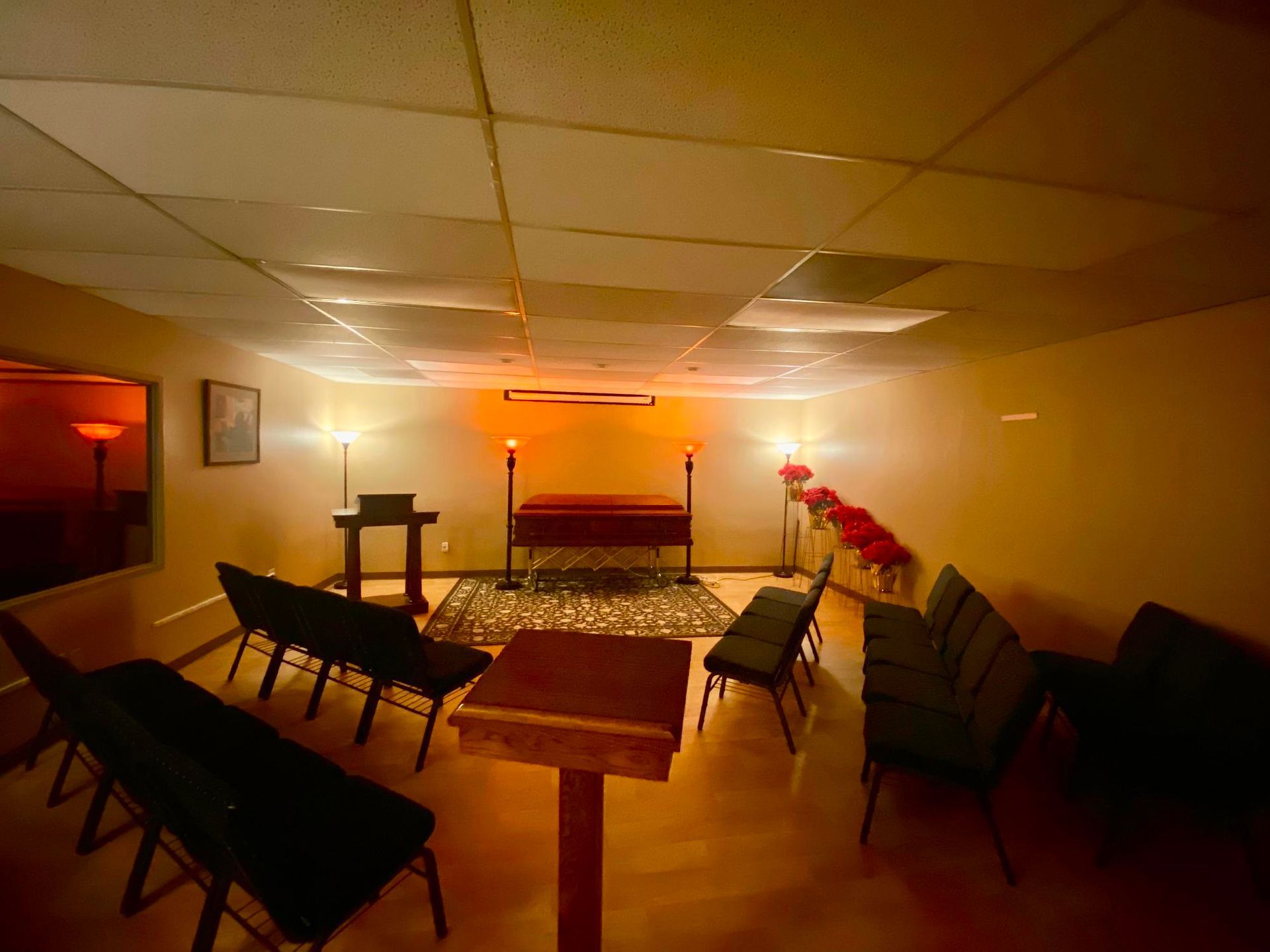 Warm-lit chapel room with rows of chairs facing a red-draped altar and a cross at the back.