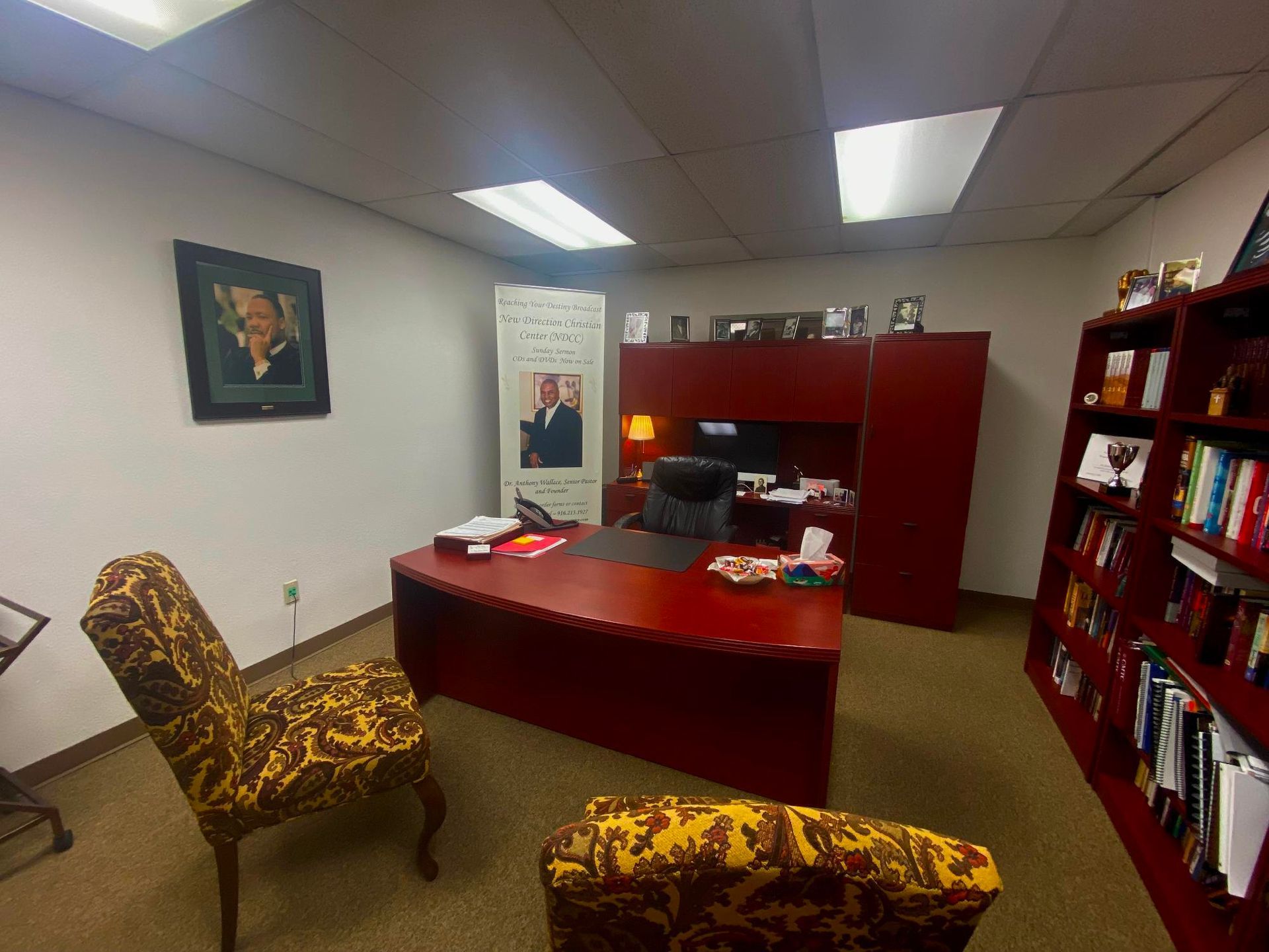 Small office with red desk and cabinets, two patterned chairs, bookshelves, and fluorescent ceiling lights.