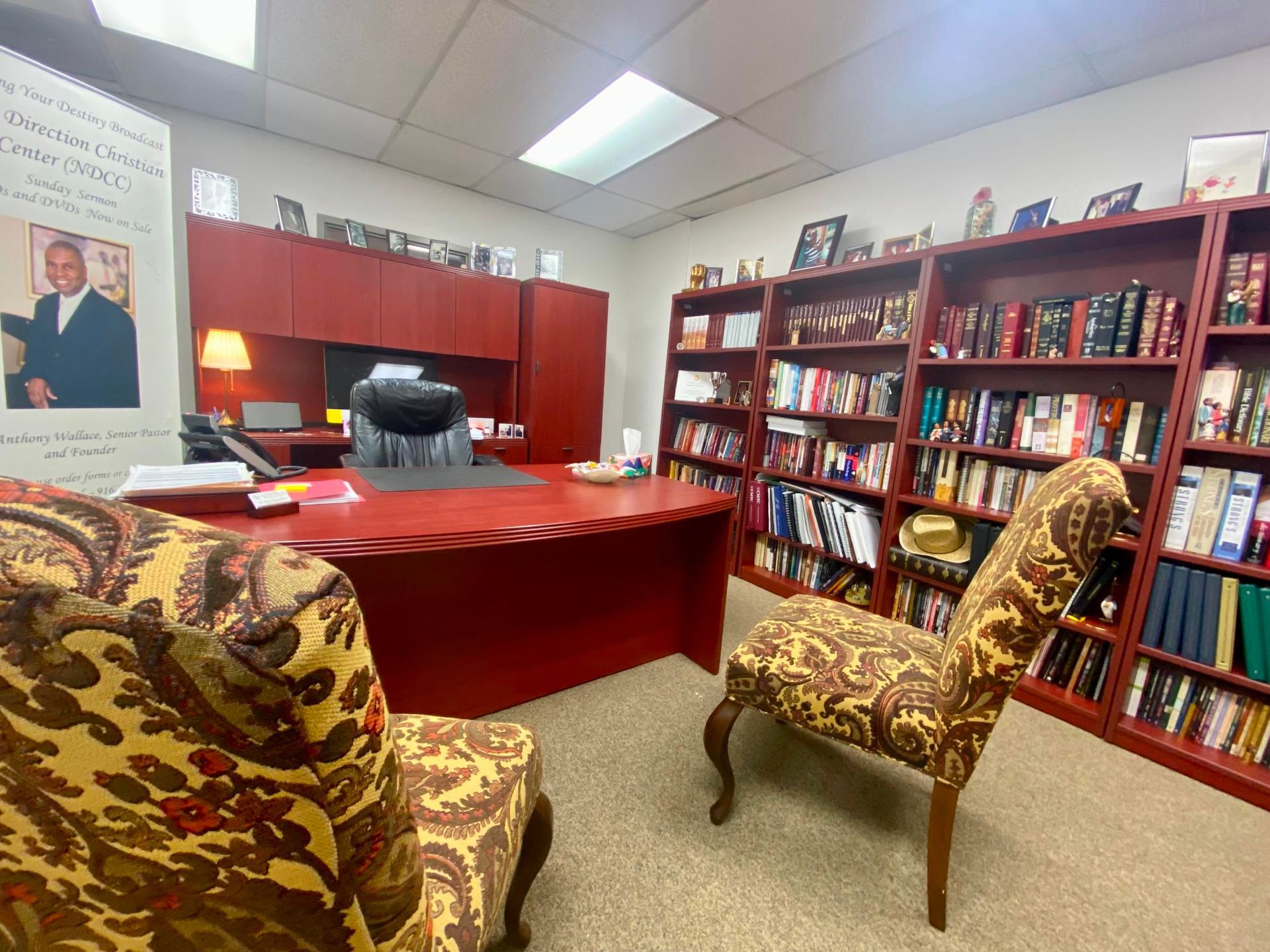 Office with red desk, bookshelves, patterned chairs, and a framed portrait on the wall.