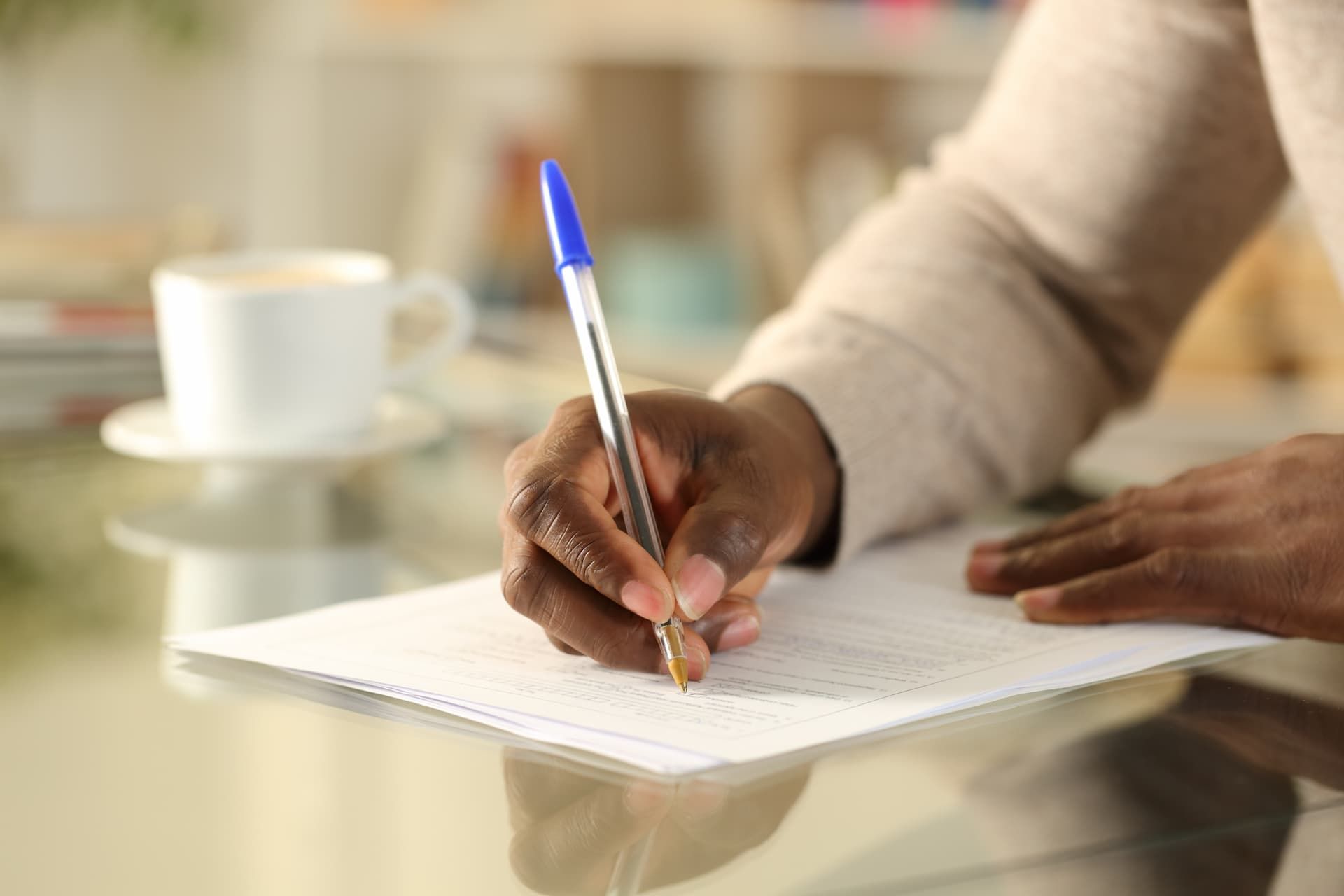 Hand writing with a blue pen on paper at a table, with a coffee cup in the background