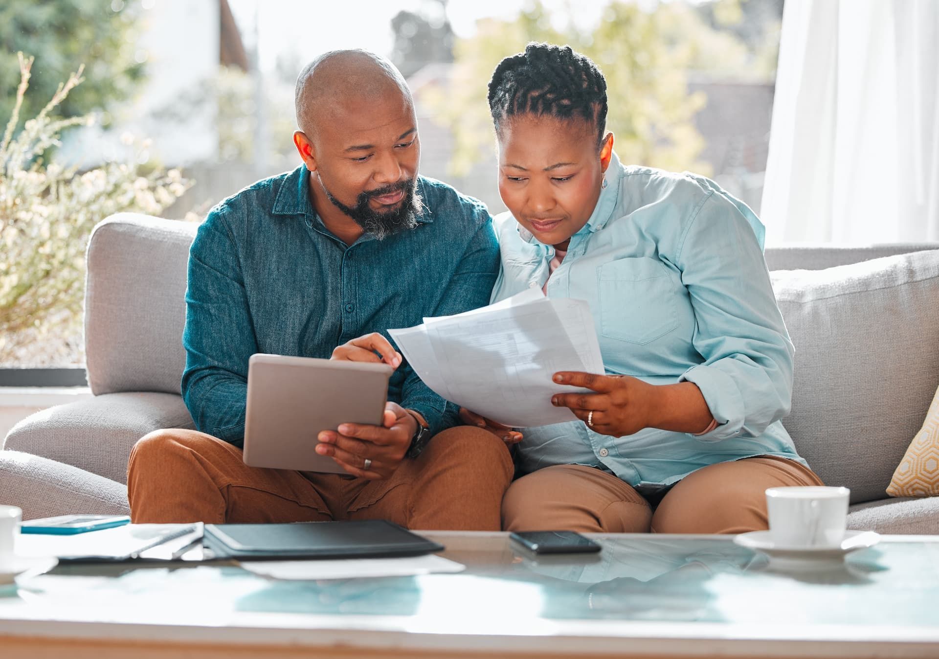 Two people sit on a couch, reviewing papers and a tablet together in a bright living room.