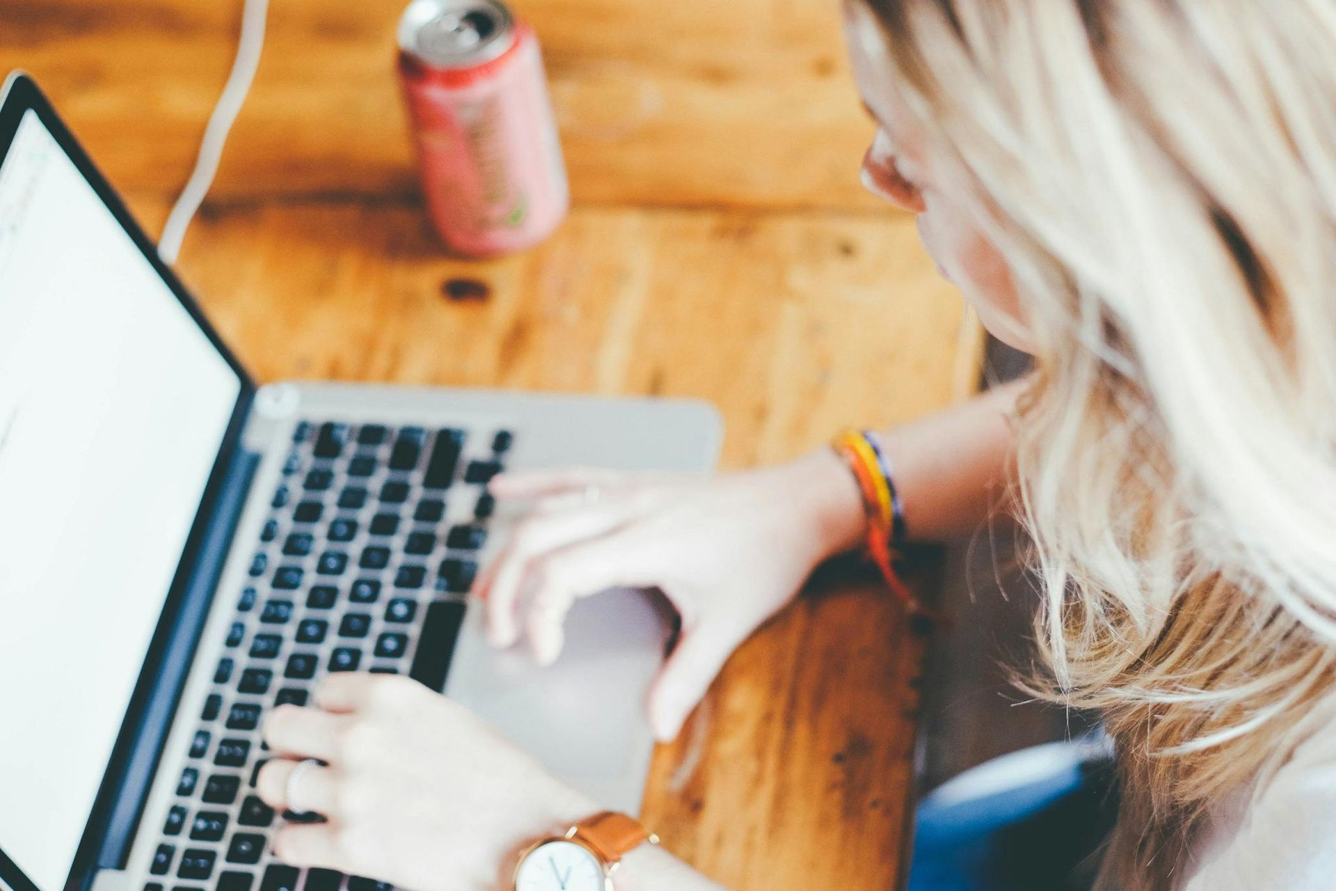 A person typing on a laptop at a wooden table with a beverage can nearby.