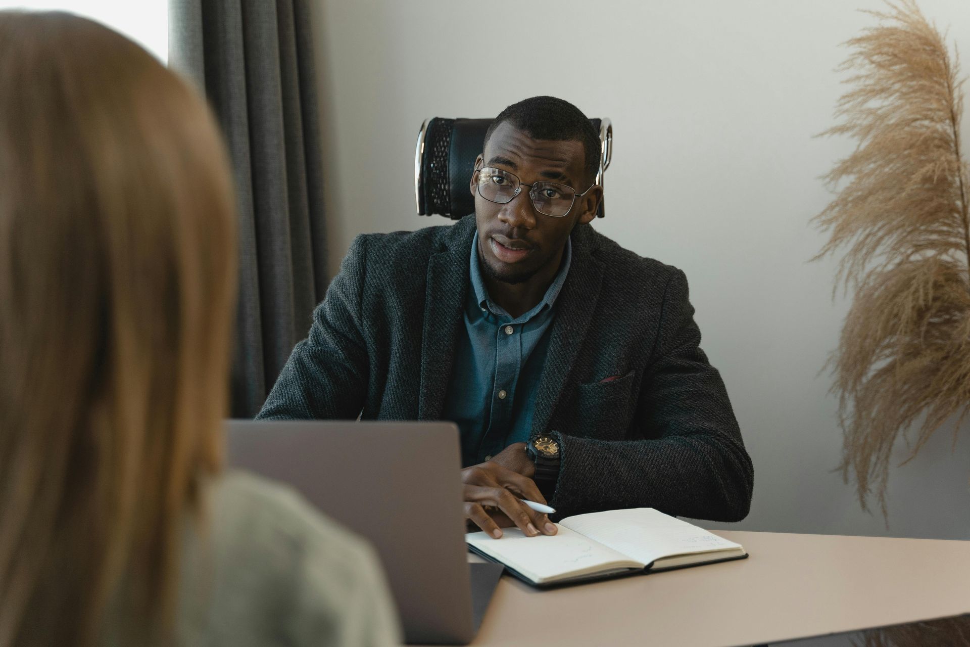 Man in glasses interviews someone at a desk, looking thoughtful. Laptop and open notebook in front.