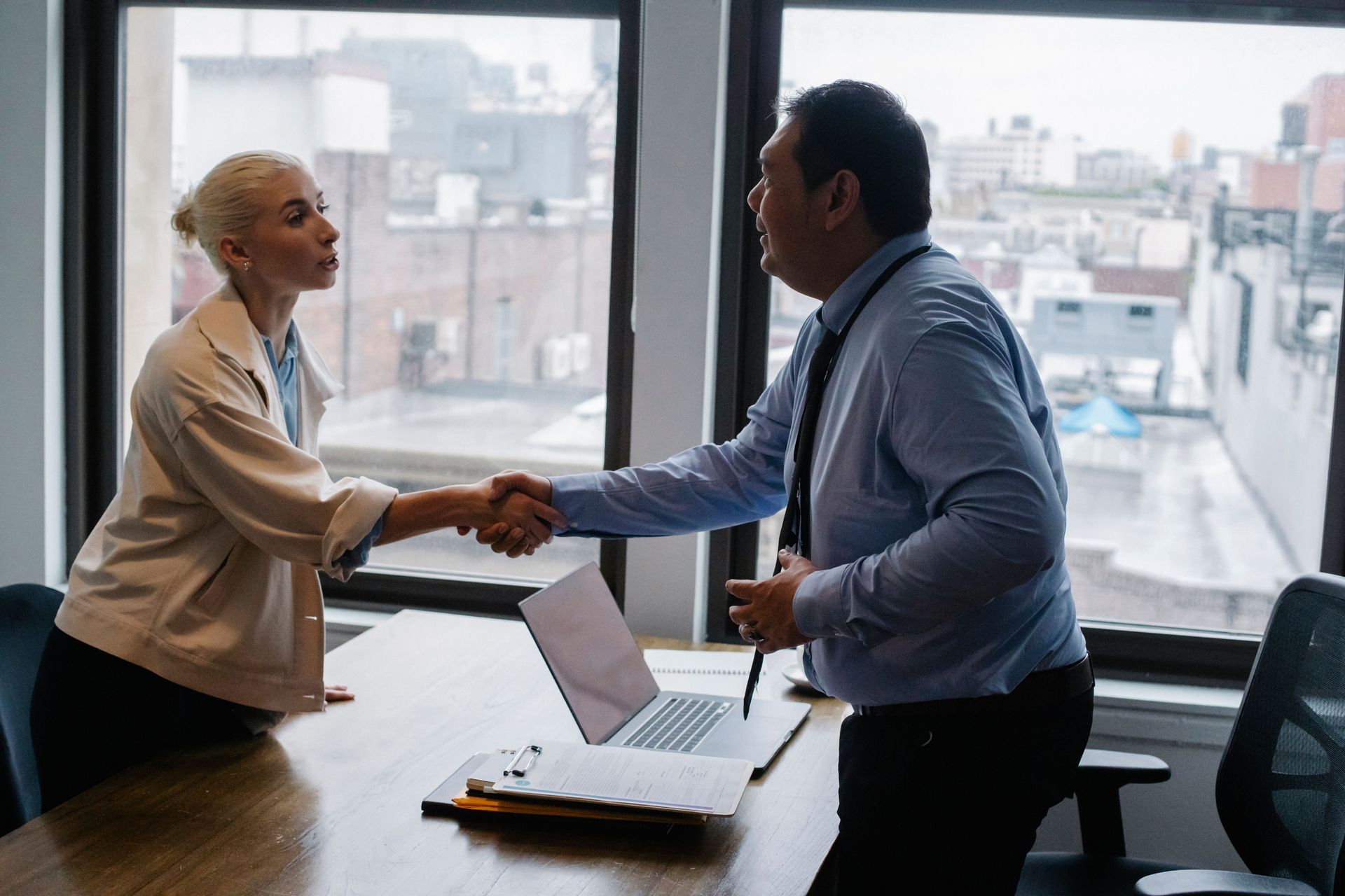 Two coworkers shake hands across a desk in a bright office with city windows.