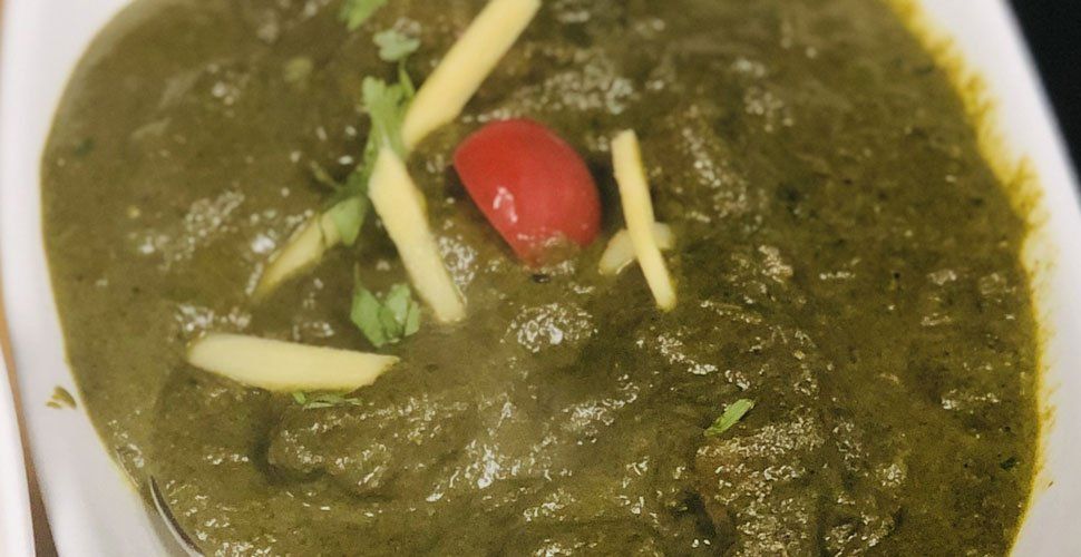Green spinach-based curry with meat, garnished with ginger and a cherry tomato, in a white bowl.