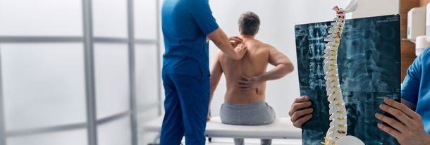 Chiropractor examining a seated patient’s back next to a spinal X-ray in a clinic