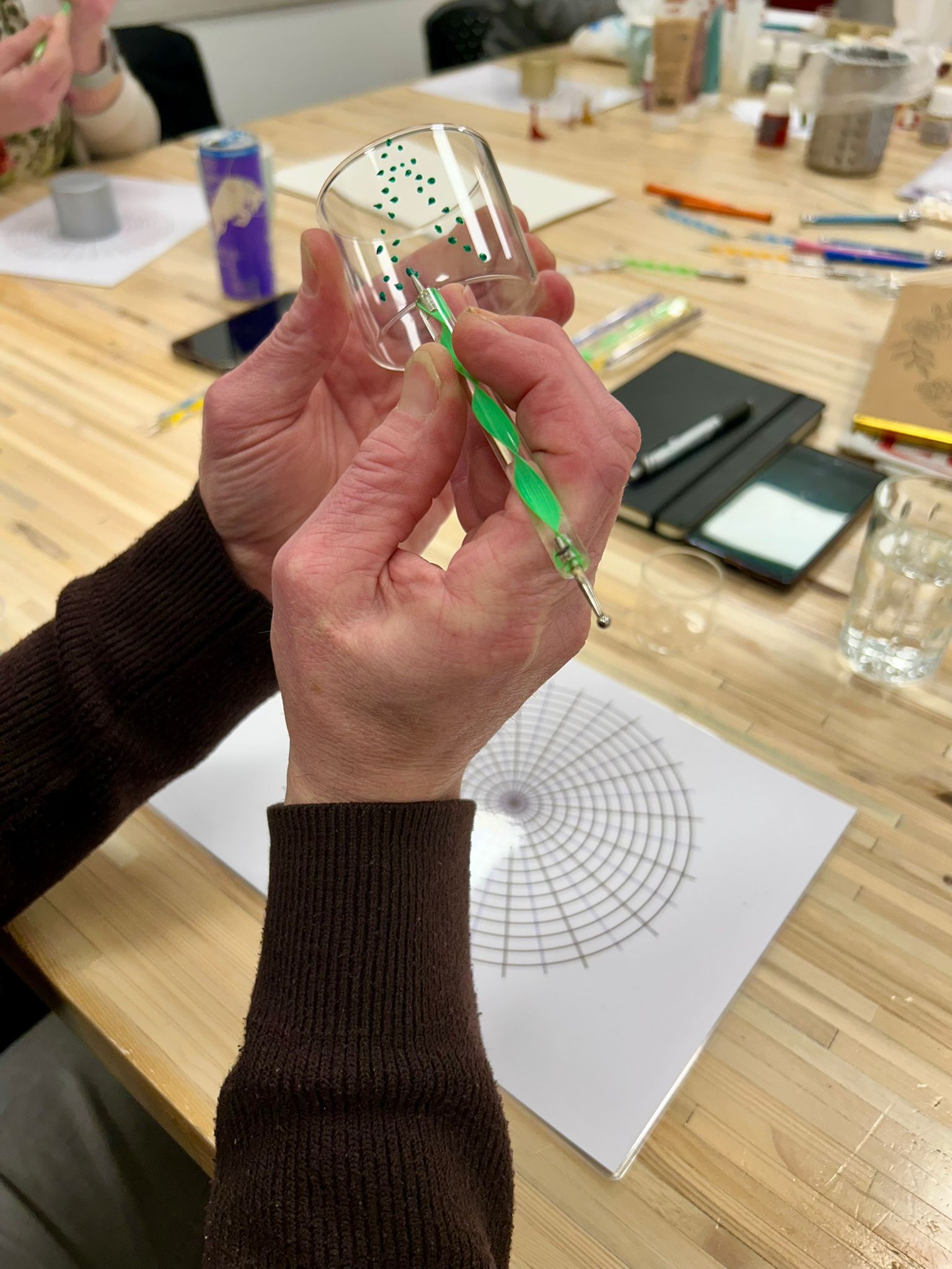 Handen versieren een glas met een groene pen, zittend aan een houten tafel met een radiaal patroon op papier.