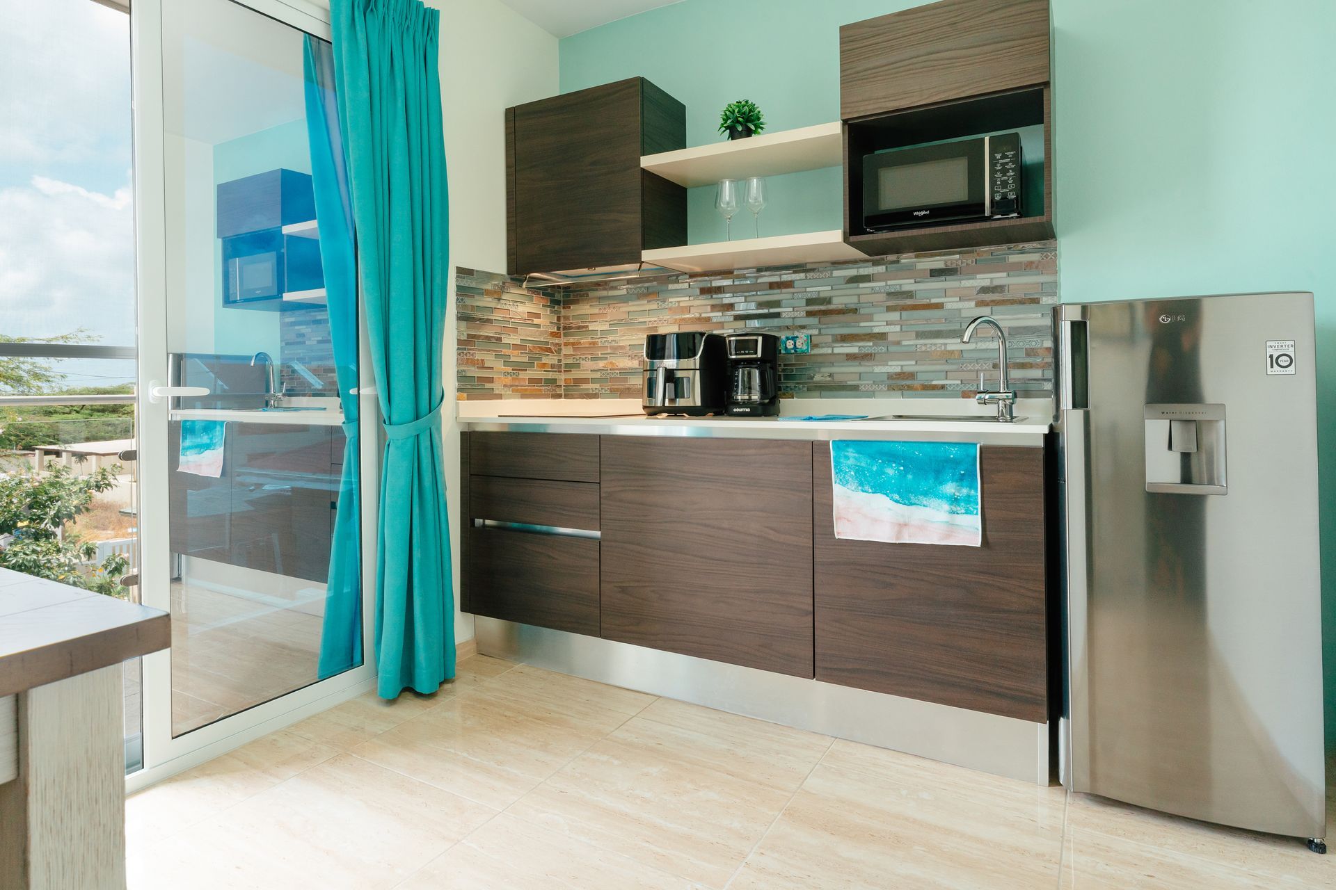 Modern kitchen with dark wood cabinets, turquoise curtains, stainless fridge, and ocean view through glass doors