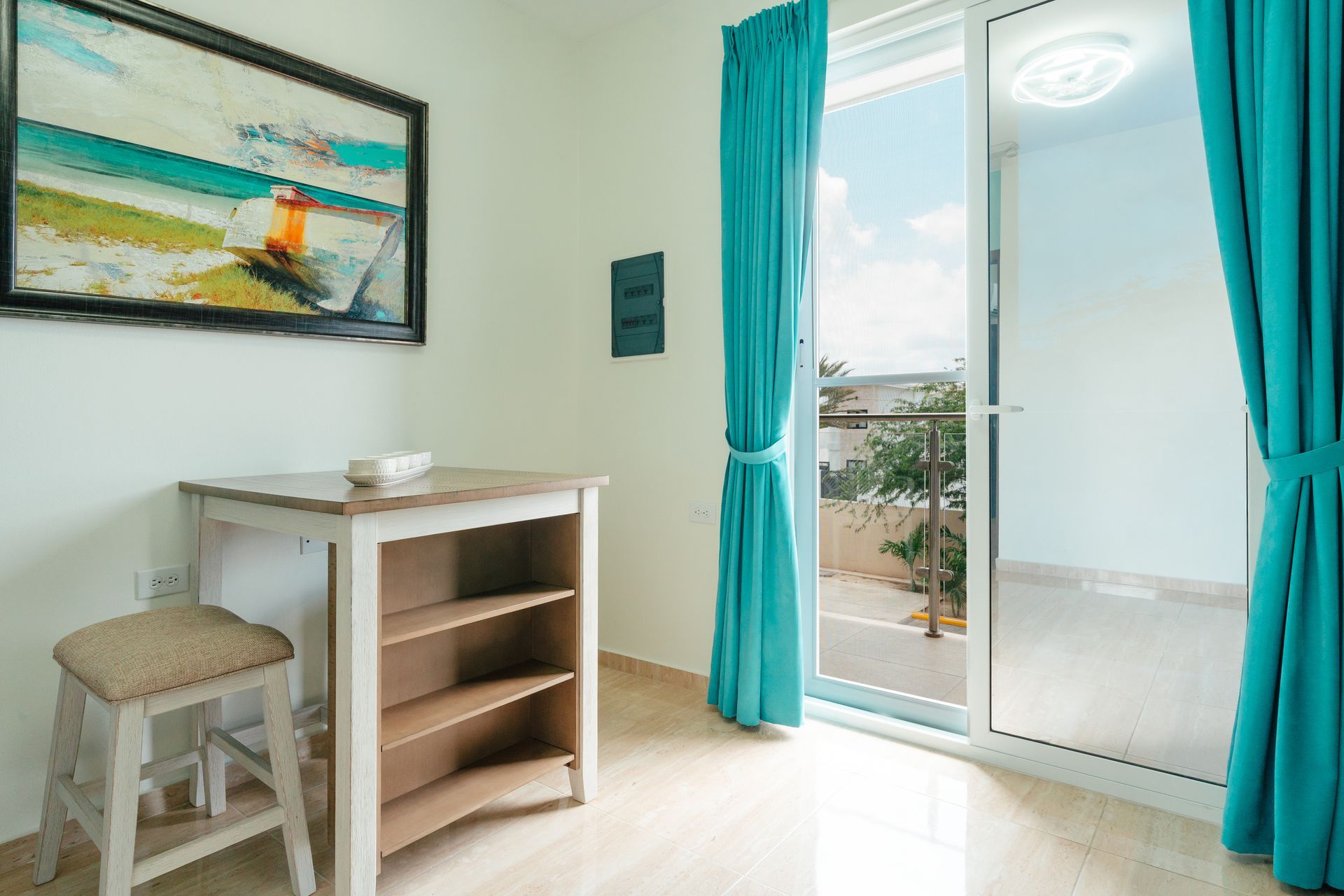 Bright room with desk, stool, wall art, and open balcony doors with turquoise curtains.