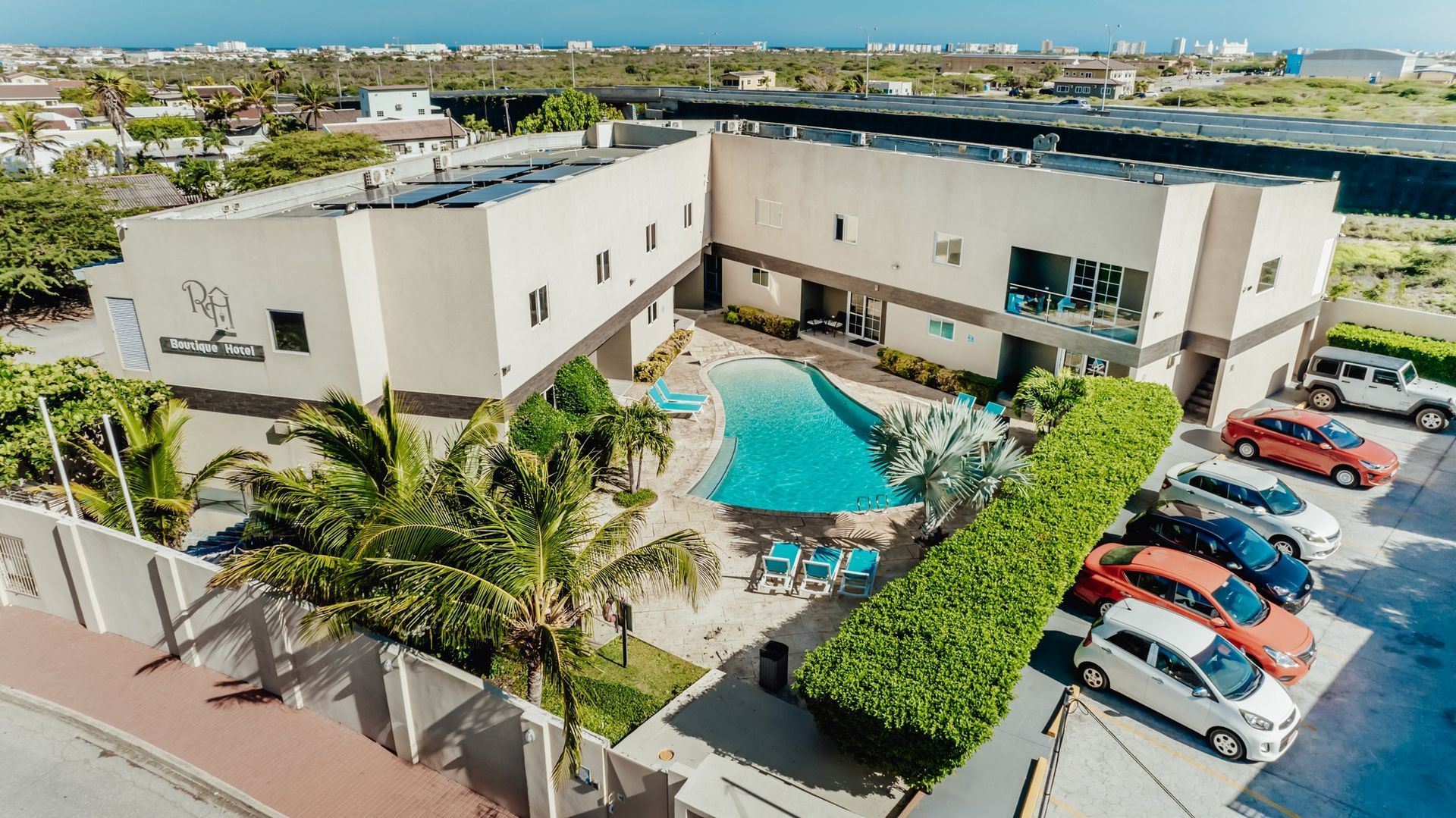 Aerial view of a hotel courtyard with a turquoise pool, palm trees, lounge chairs, and parked cars nearby