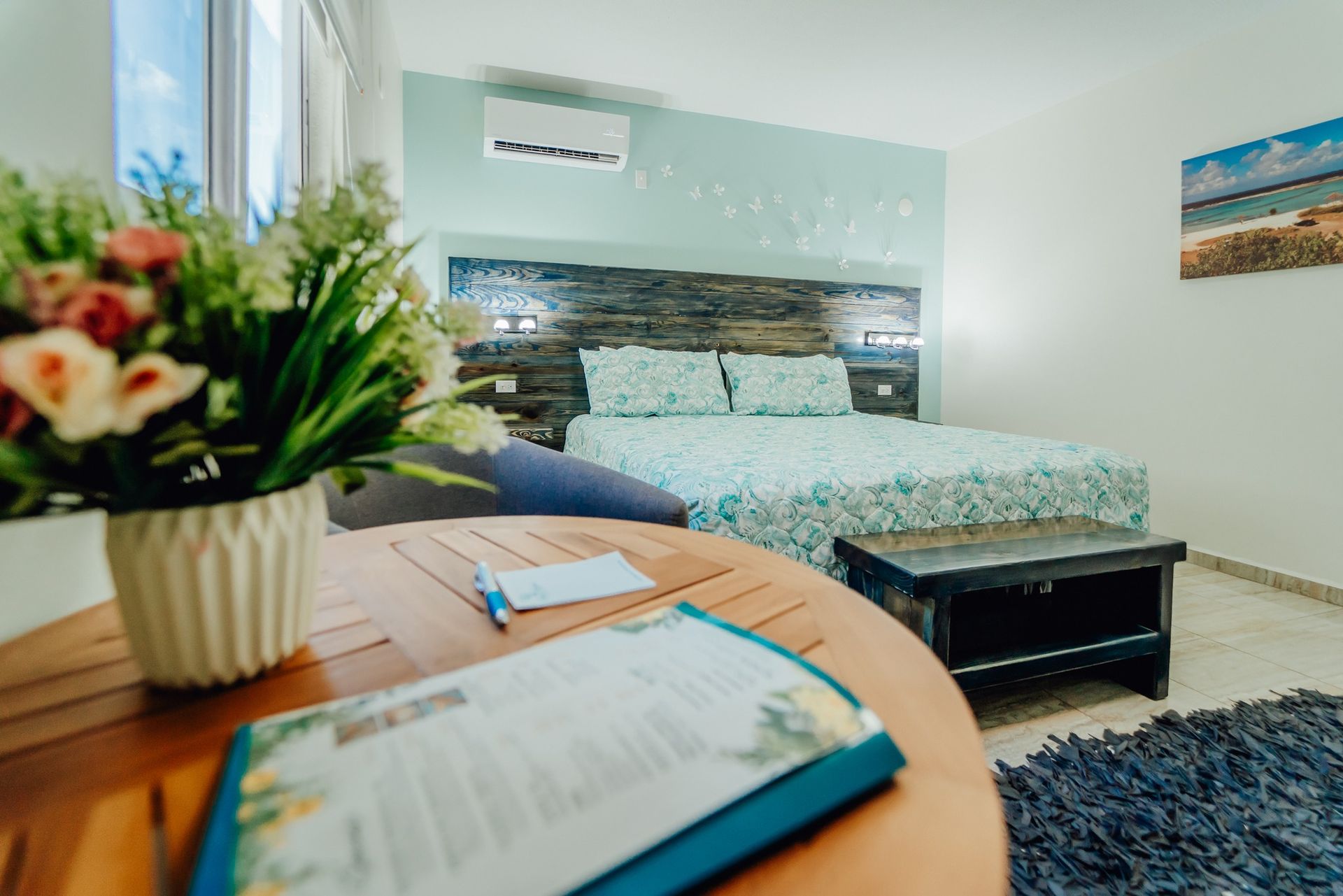 Bright bedroom with a neatly made bed, wooden table, flowers, and a book in the foreground